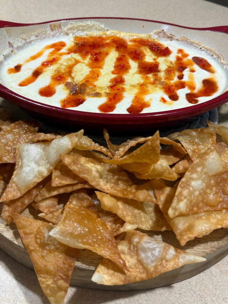 A red dish filled with creamy, melted cheese dip topped with a drizzle of red sauce, served on a wooden board with crispy fried wonton chips.