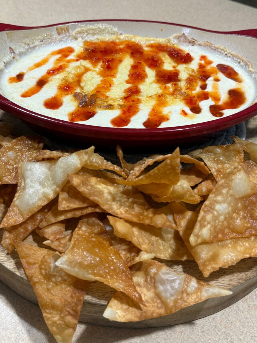 A red dish filled with creamy, melted cheese dip topped with a drizzle of red sauce, served on a wooden board with crispy fried wonton chips.
