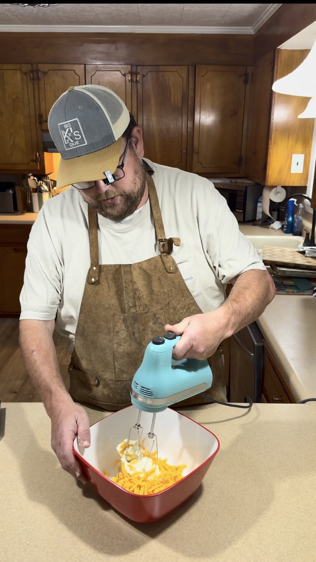 A man in a brown apron and baseball cap uses a blue hand mixer to blend ingredients in a red and white mixing bowl on a kitchen counter. Wooden cabinets and various kitchen items are visible in the background.