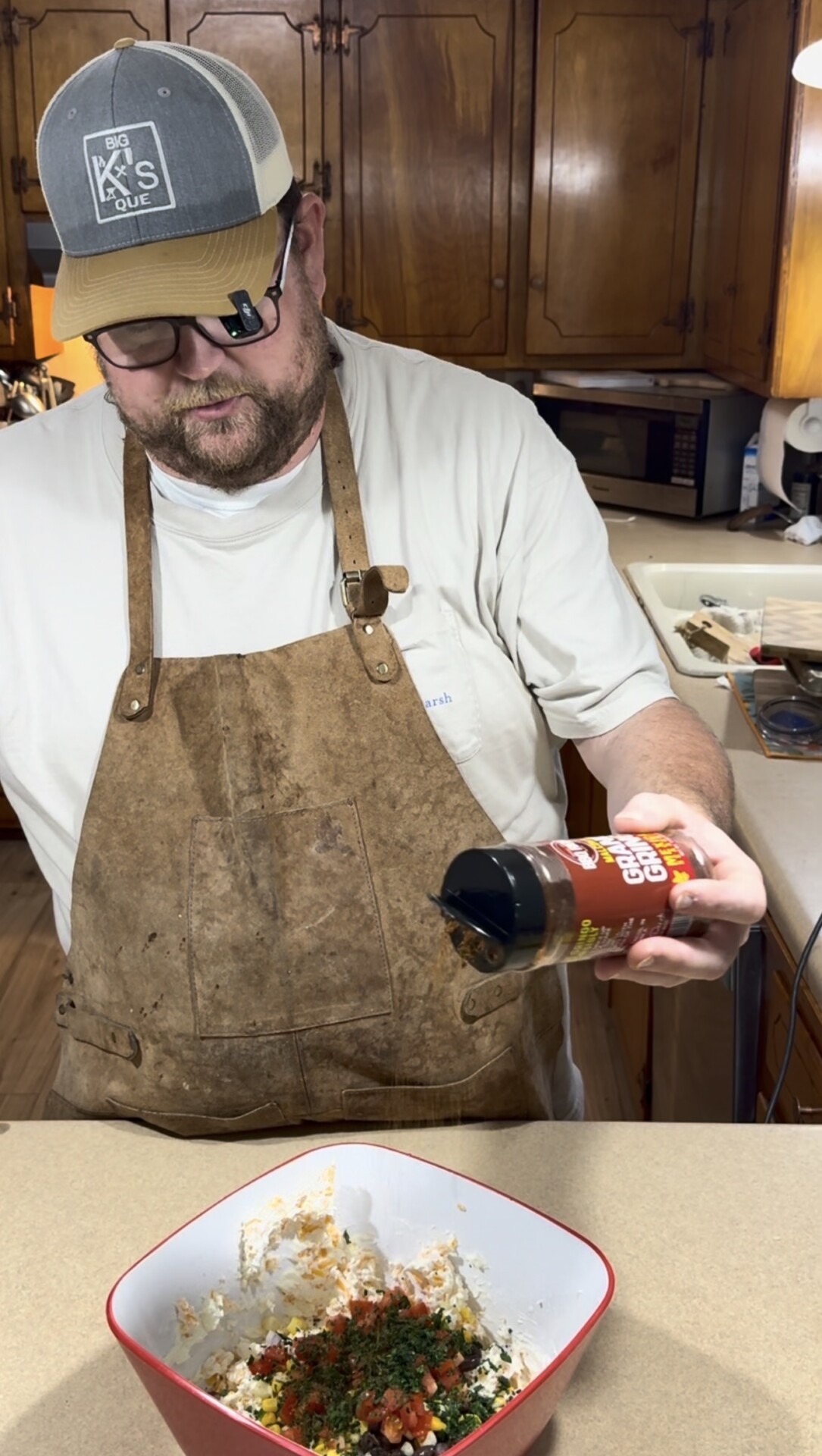 A man wearing a brown apron and beige hat sprinkles seasoning from a bottle into a bowl of mixed ingredients on a kitchen counter. Wooden cabinets and a microwave are visible in the background.