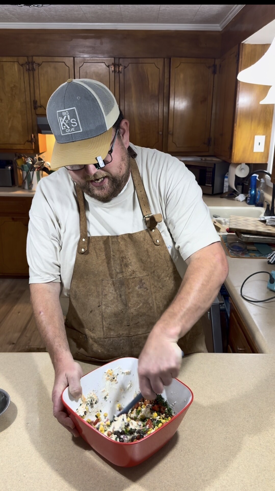 A man wearing a tan hat, glasses, a white shirt, and a brown apron is mixing ingredients in a red and white bowl on a kitchen counter with wooden cabinets in the background.