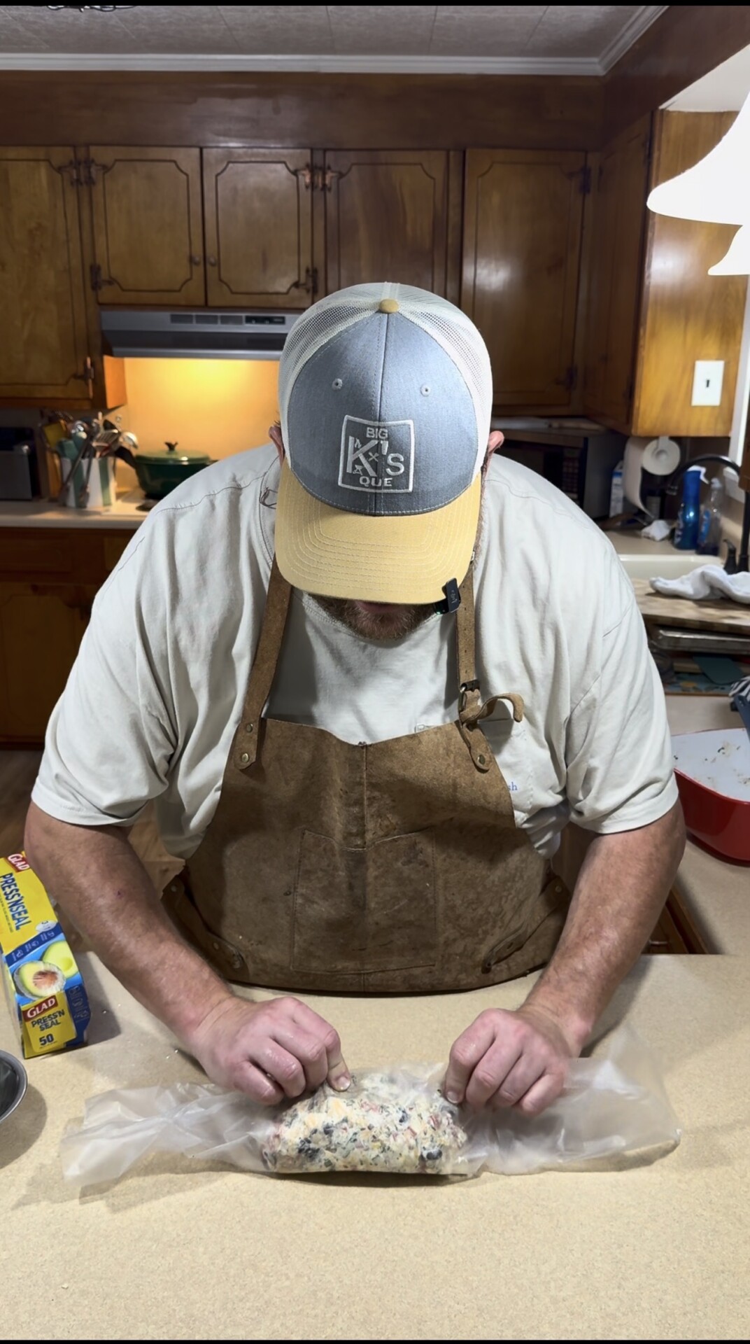 A man wearing a cap, apron, and t-shirt is rolling a mixture in parchment paper on a kitchen counter, with plastic wrap and utensils nearby in a warm-lit kitchen.