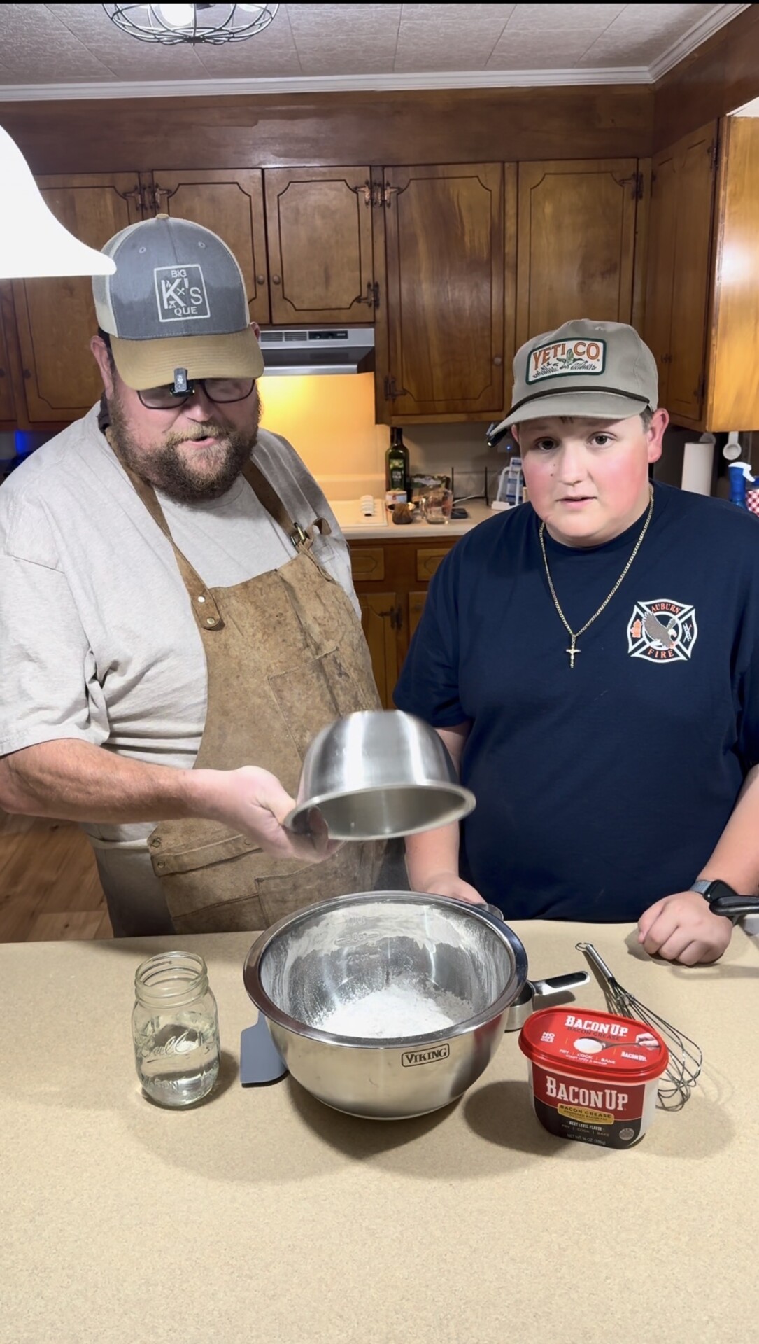 Two people stand in a kitchen, preparing to cook. The person on the left wears an apron and holds a metal bowl over a larger mixing bowl with flour. The person on the right stands next to him. Ingredients and utensils are on the counter.