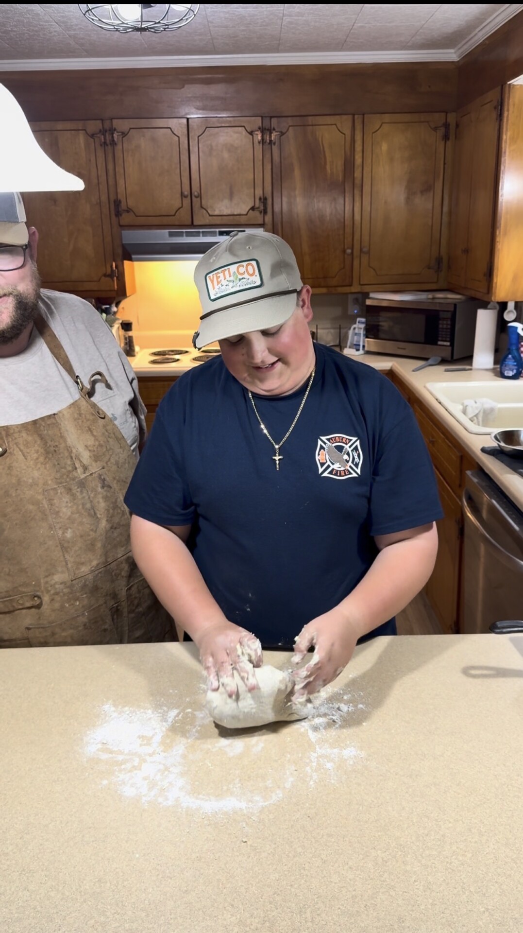 A boy wearing a hat and dark t-shirt kneads dough on a floured kitchen counter, while a bearded man in an apron stands nearby. The kitchen has wooden cabinets and warm lighting.