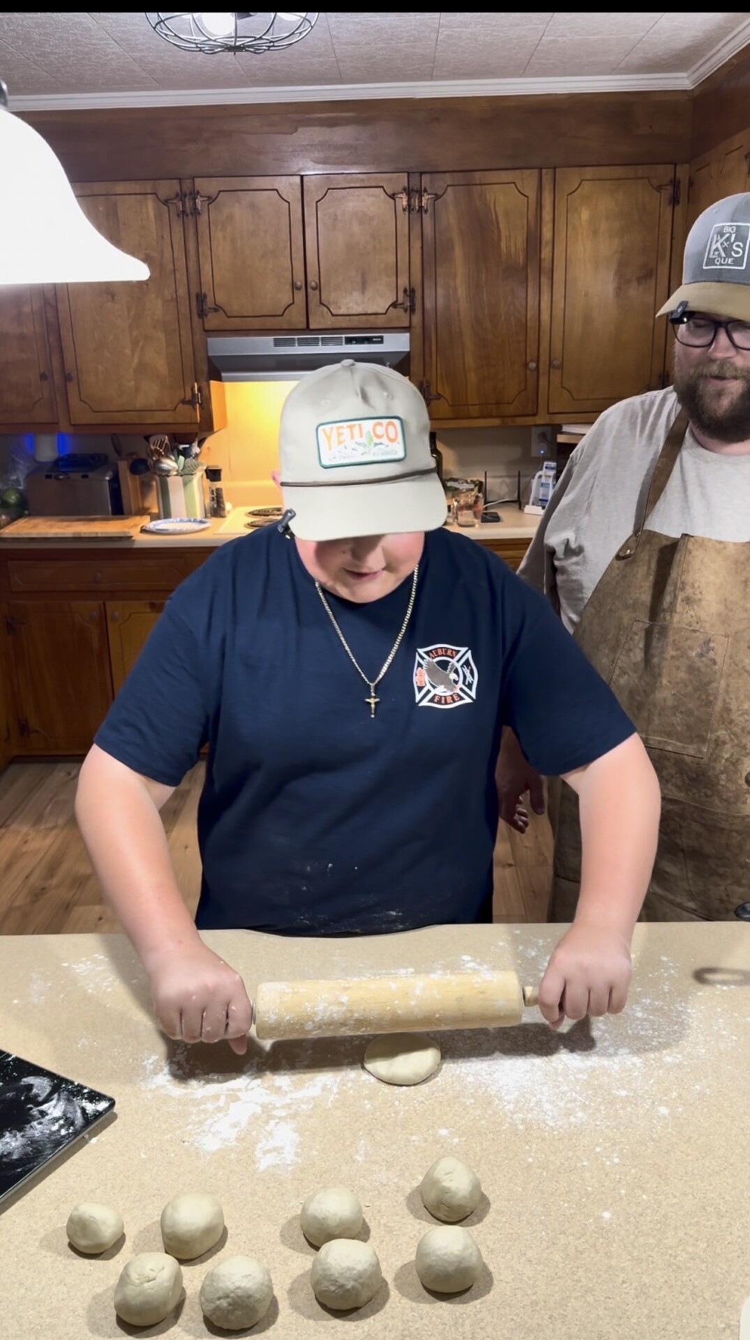 A young person rolls dough with a rolling pin on a floured countertop, while a man in an apron and hat stands beside them in a kitchen with wooden cabinets. Several dough balls are visible on the counter.