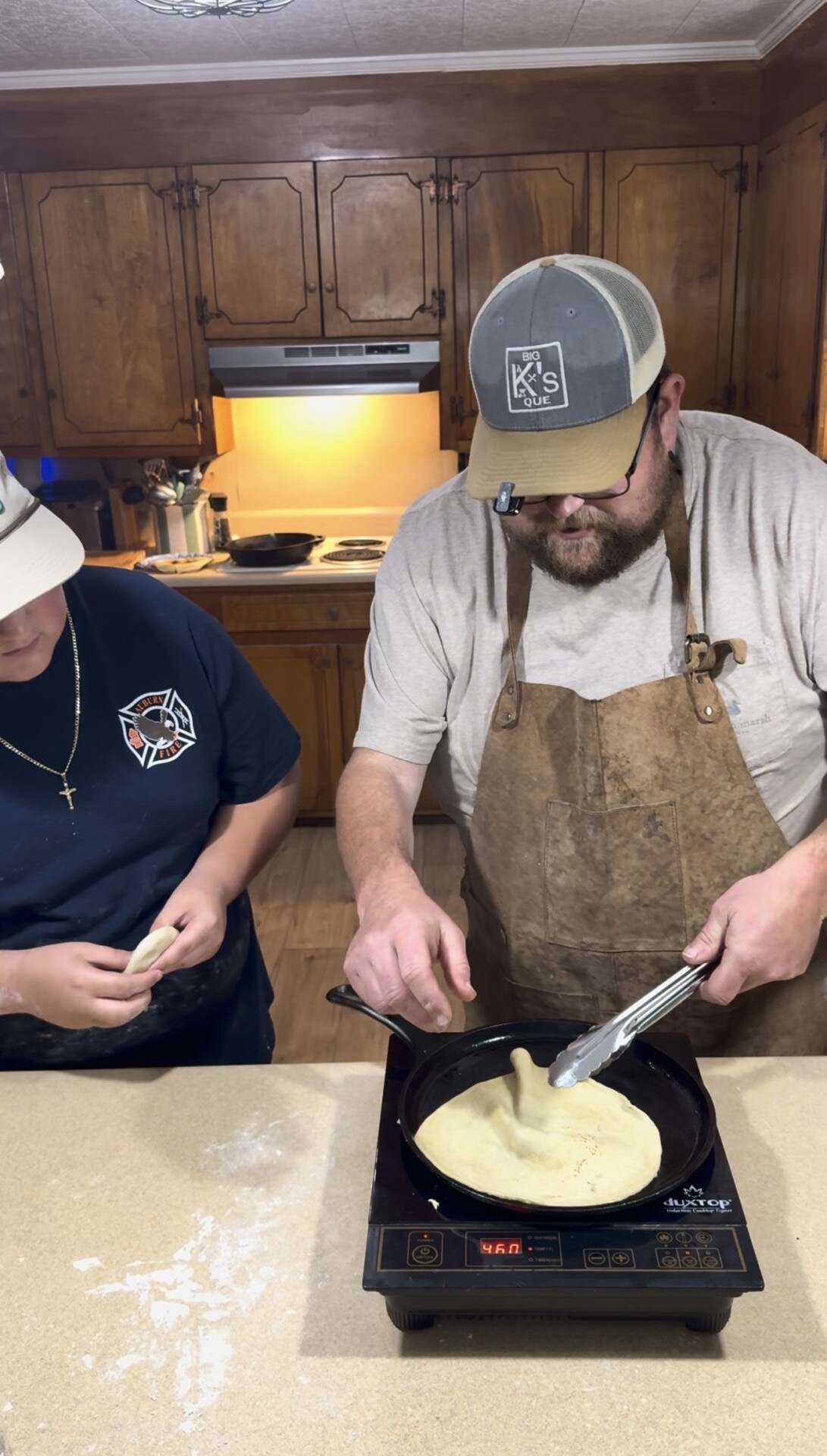 Two people in a kitchen prepare dough; one places a flat piece into a skillet on an induction cooktop, while the other shapes dough. Both wear hats, and one wears an apron. Wooden cabinets and warm lighting are in the background.