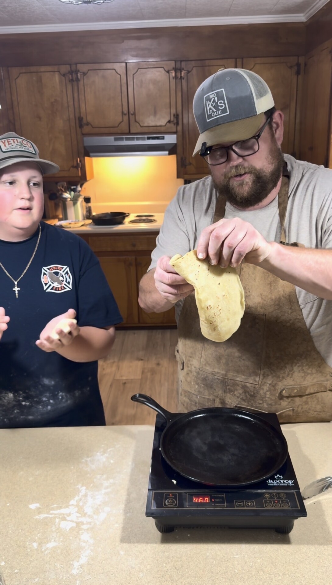 A man wearing an apron and cap stretches dough over a skillet on an electric stove, while a child beside him claps flour from their hands in a kitchen with wooden cabinets.