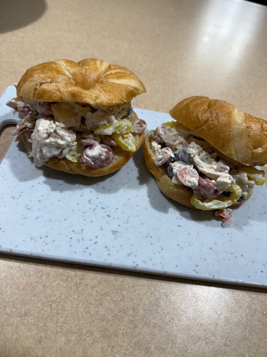 Two croissant sandwiches filled with a creamy mixture of chicken, diced vegetables, sliced olives, and banana peppers are placed on a white speckled cutting board on a tan countertop.