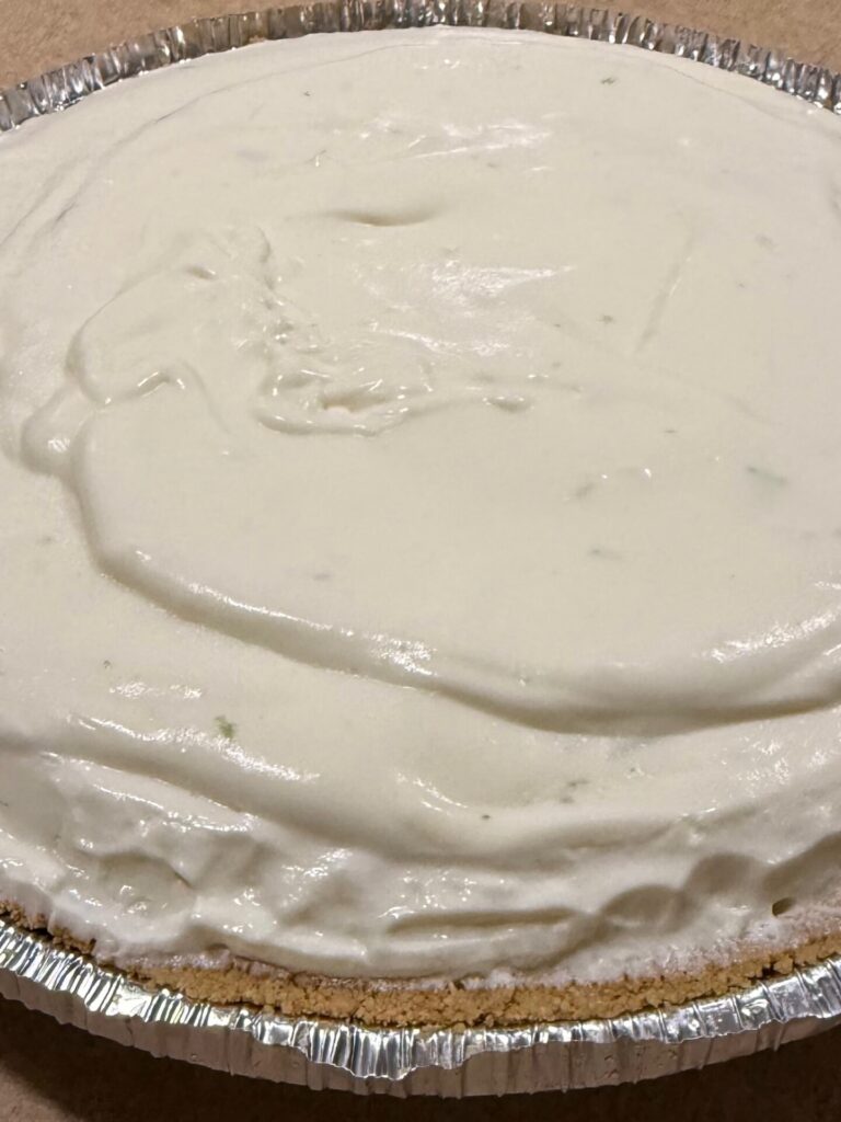 A close-up of a creamy, white pie with a graham cracker crust in an aluminum pan, sitting on a flat surface. The pie appears to be unbaked or a no-bake dessert.