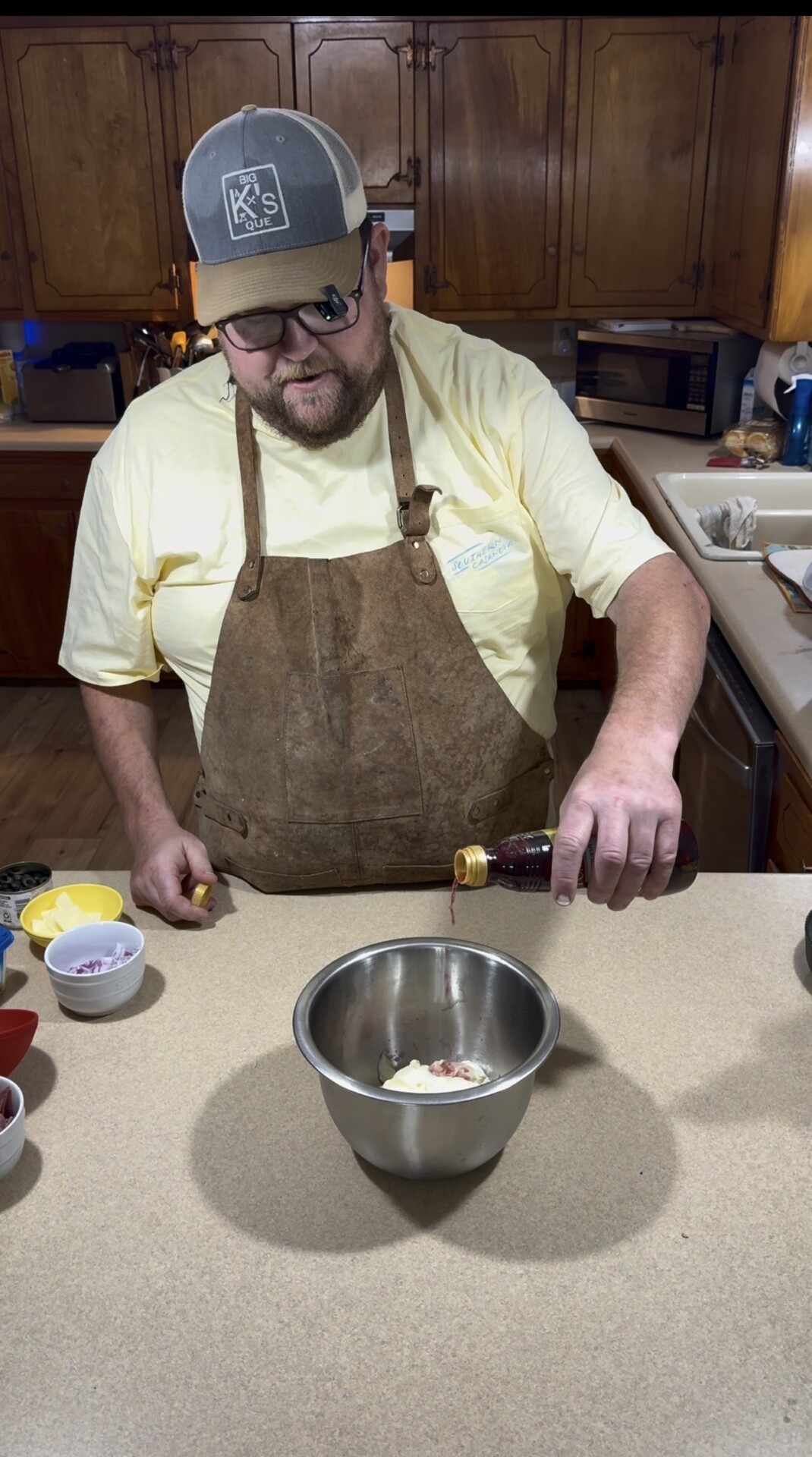 A man wearing an apron and cap pours a liquid into a metal bowl on a kitchen counter, surrounded by small bowls with ingredients.