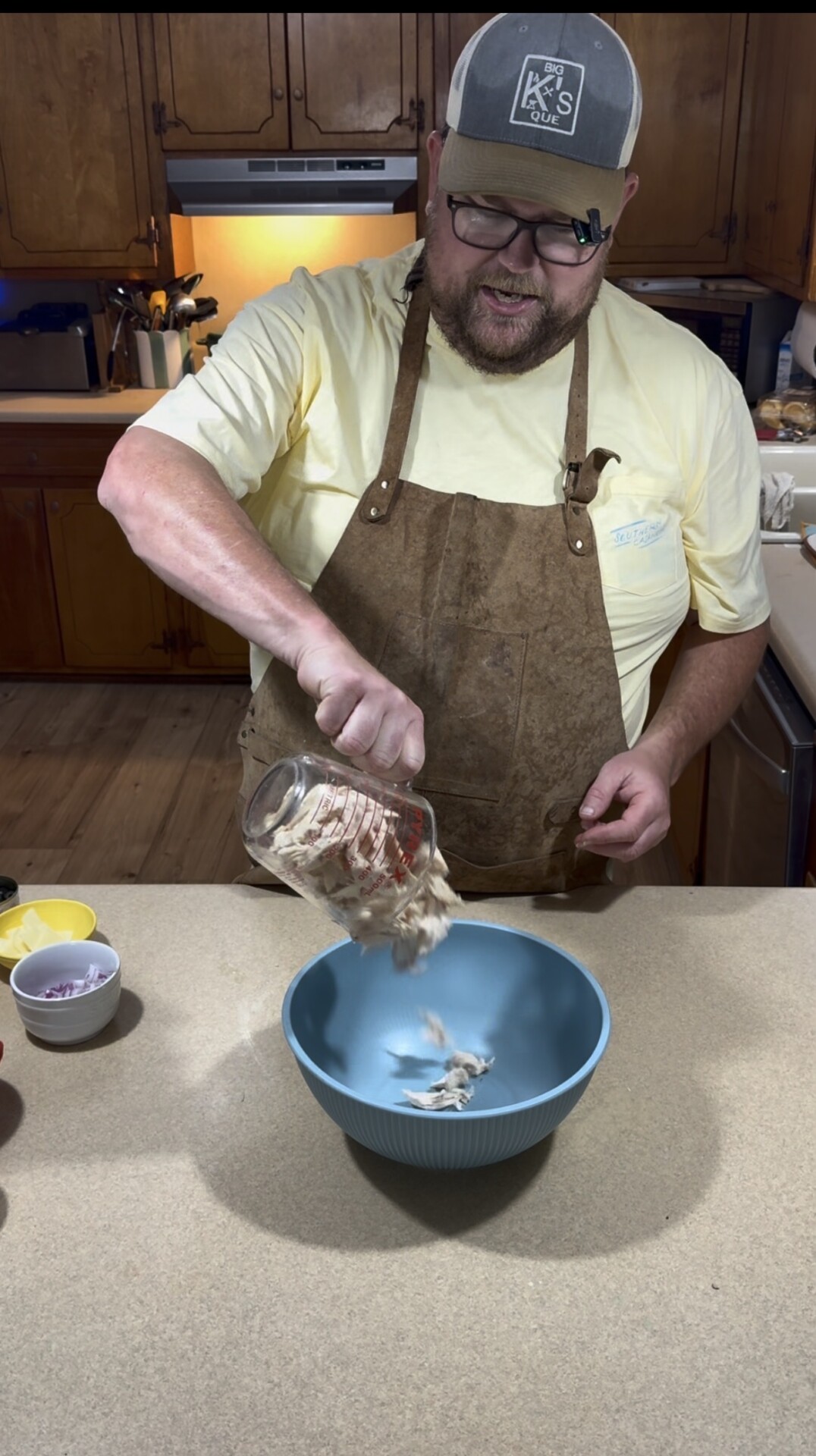 A man in an apron and hat pours chopped onions from a glass measuring cup into a blue mixing bowl on a kitchen counter.