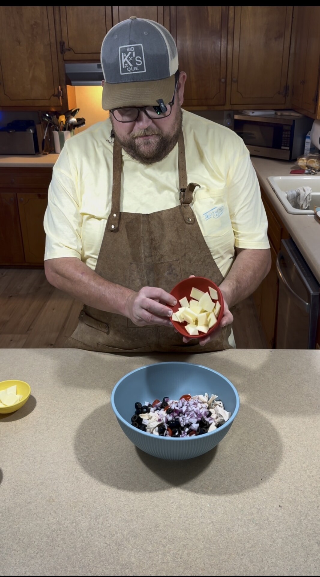 A man in a yellow shirt and brown apron adds cubed butter from a red bowl into a blue mixing bowl on a kitchen counter, preparing ingredients.