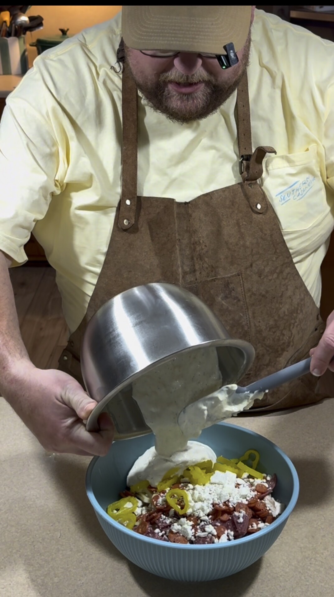 A person wearing a brown apron and yellow shirt pours creamy dressing from a metal bowl onto a salad topped with bacon, feta cheese, and pepper slices in a blue bowl.
