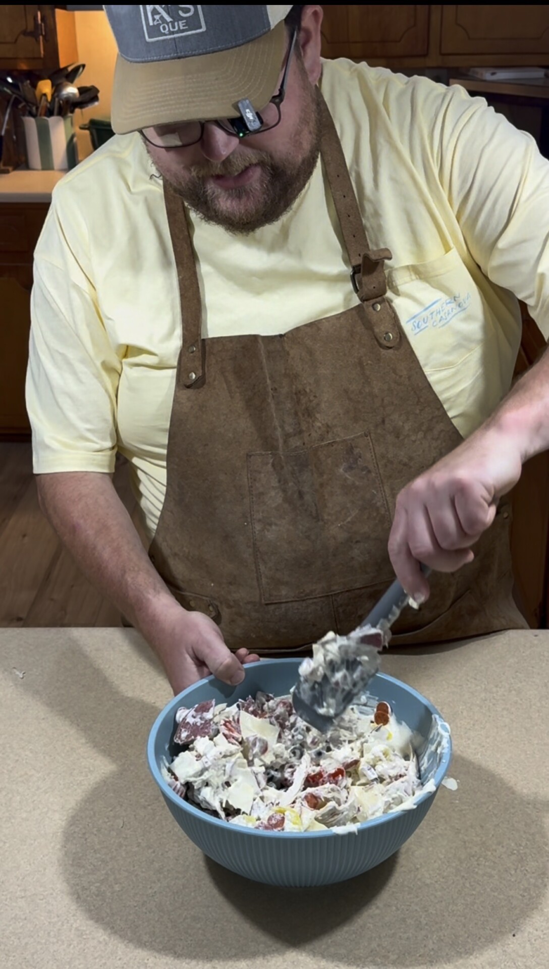 A man wearing a brown apron and yellow shirt stands in a kitchen, mixing a large bowl of salad with a serving spoon.
