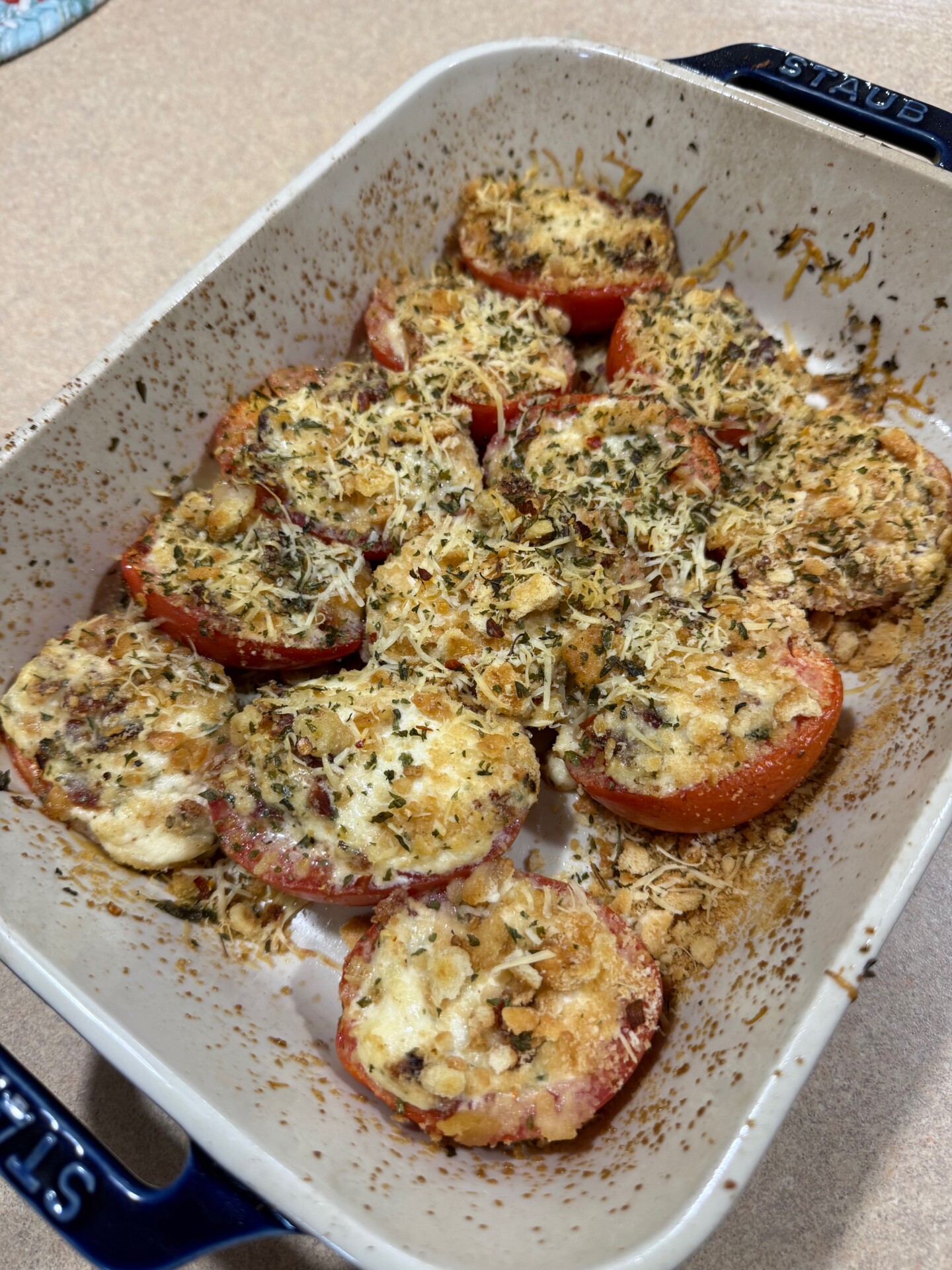 Sliced tomatoes in a baking dish topped with breadcrumbs, herbs, and melted cheese, baked until golden brown. The dish is on a beige countertop.