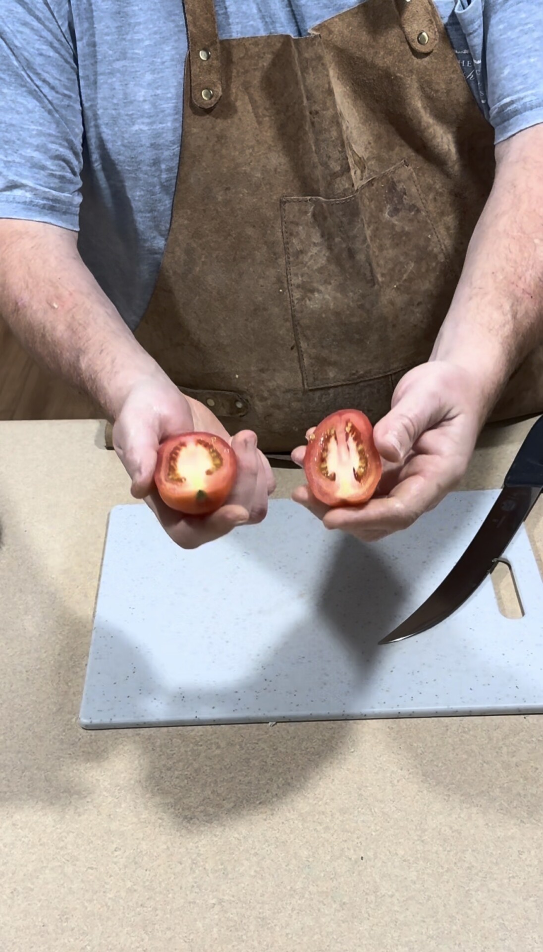 A person wearing a brown apron holds a tomato cut in half over a cutting board, with a knife visible to the right.