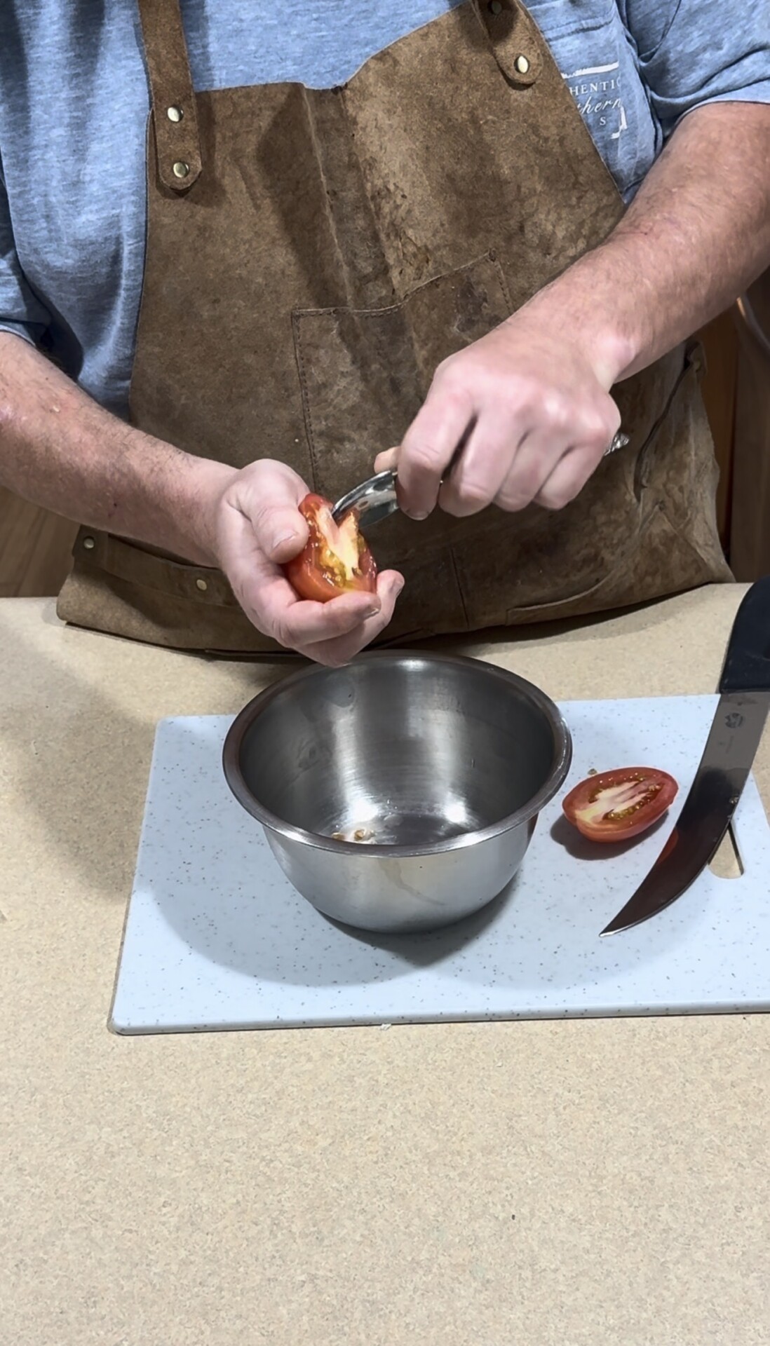 A person in a brown apron uses a spoon to scoop seeds from a halved tomato over a metal bowl. A knife and another tomato half are on a cutting board nearby.