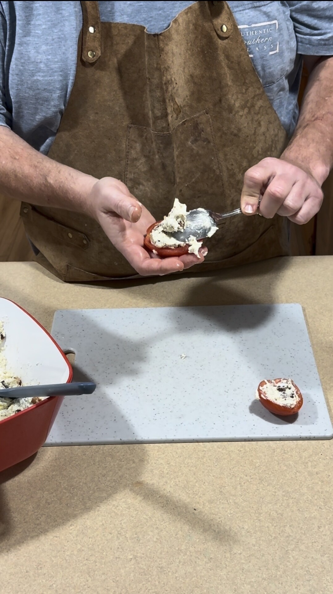 A person wearing a brown apron is filling a halved tomato with a creamy stuffing mixture using a spoon, while standing at a kitchen counter with a cutting board and mixing bowl nearby.