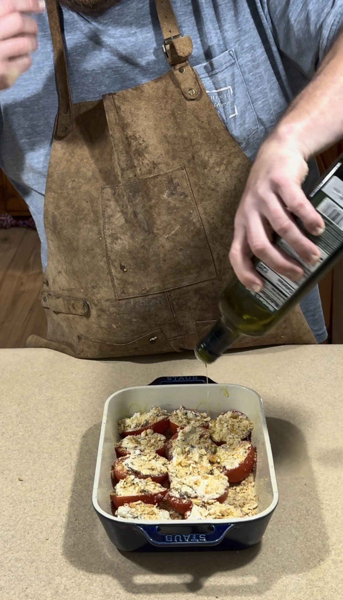 A person in a brown apron drizzles olive oil over stuffed tomatoes in a white baking dish on a kitchen counter.