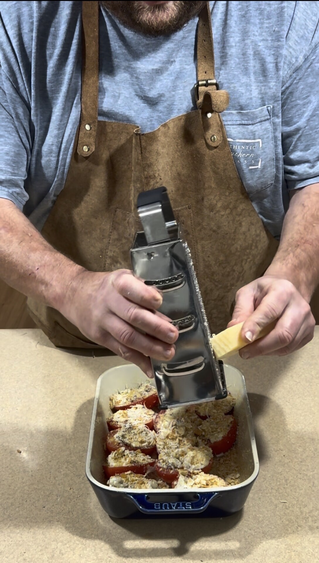 A person wearing a brown apron grates cheese over a baking dish filled with stuffed red bell peppers on a kitchen counter.