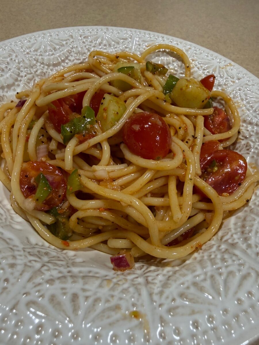 A white plate with spaghetti mixed with cherry tomatoes, chopped green peppers, onions, and herbs, sitting on a beige surface.