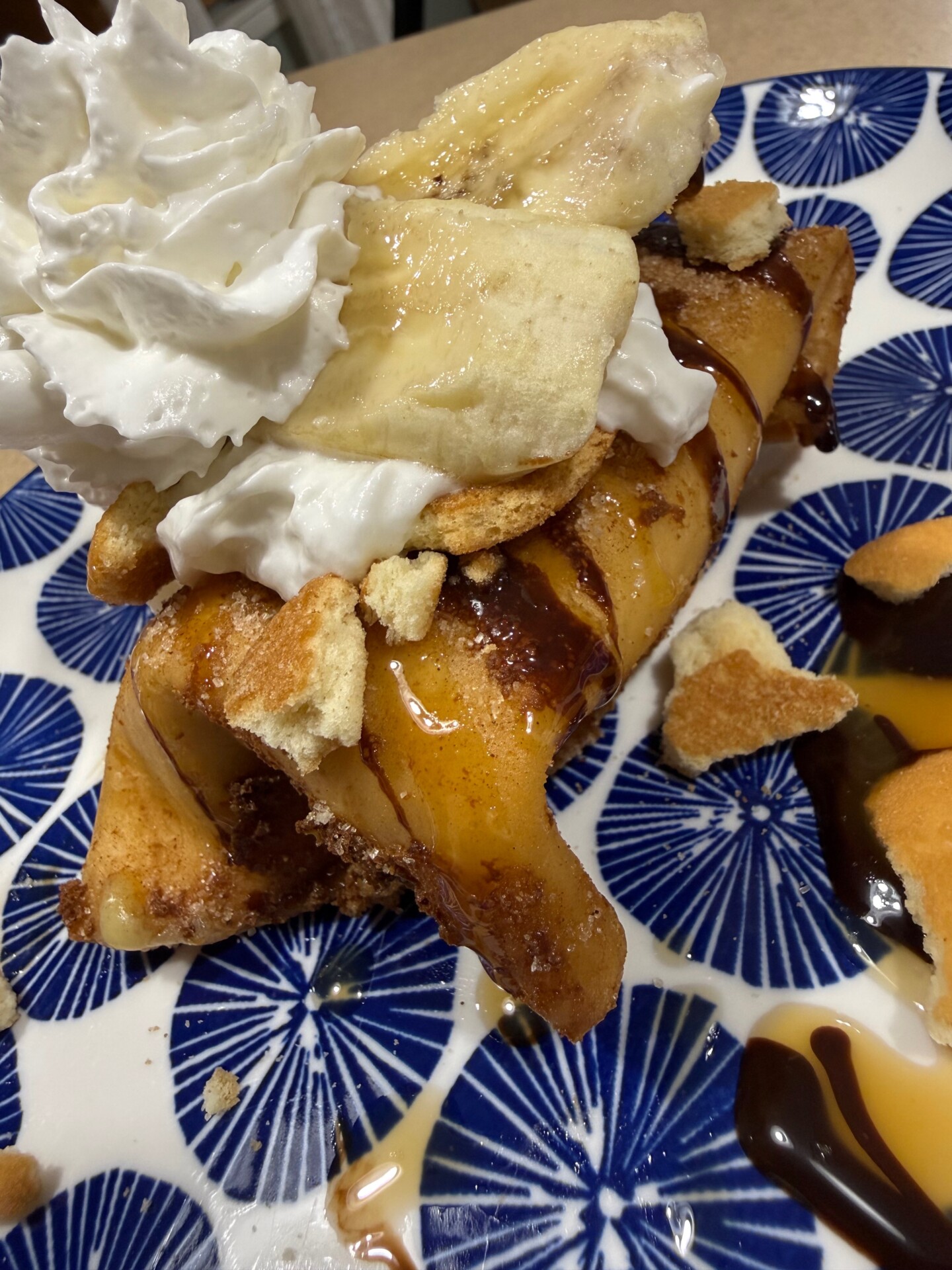 A dessert crepe topped with banana slices, whipped cream, chocolate and caramel sauce, and crumbled cookies, served on a blue and white patterned plate.