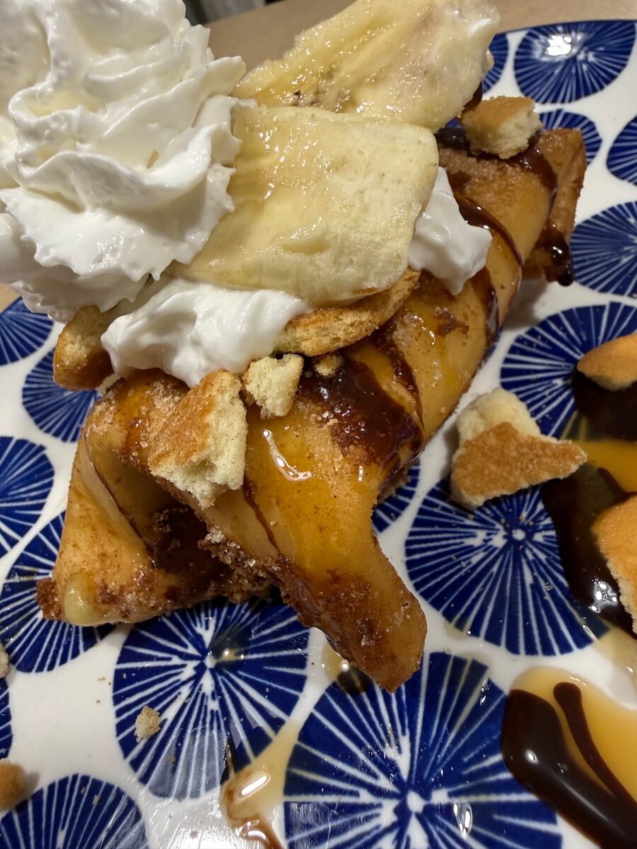 A close-up of a dessert crepe topped with whipped cream, banana slices, vanilla wafers, chocolate, and caramel sauce on a blue patterned plate.