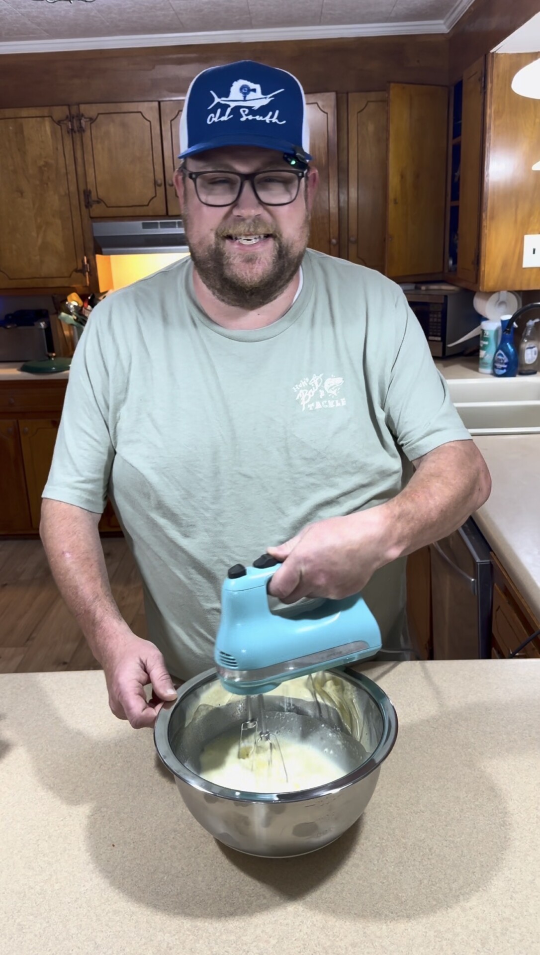 A person in glasses and a cap uses a blue hand mixer to blend ingredients in a metal bowl in a kitchen with wooden cabinets.