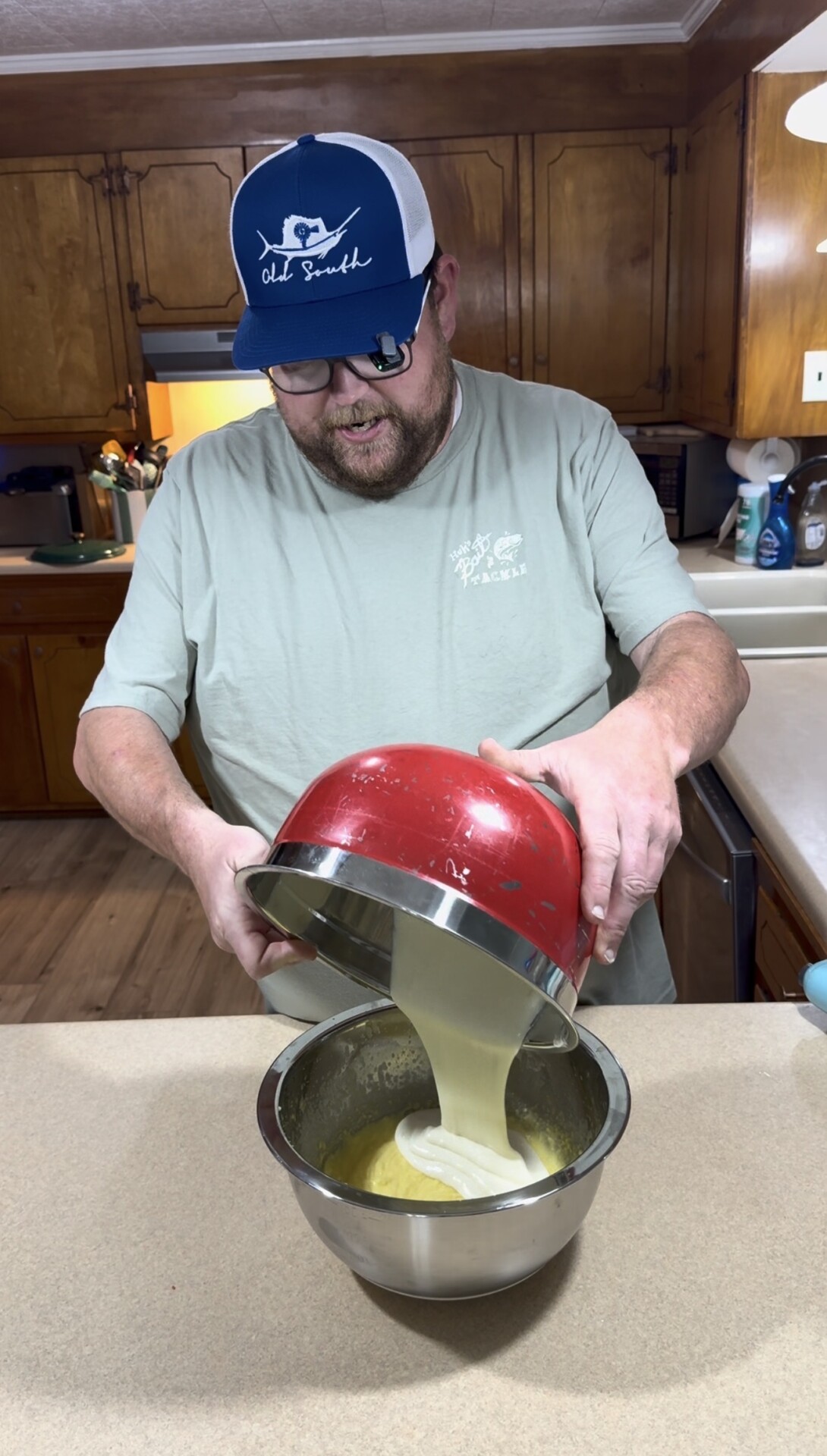 A man wearing a cap and glasses pours batter from a red bowl into a metal mixing bowl in a kitchen with wooden cabinets.