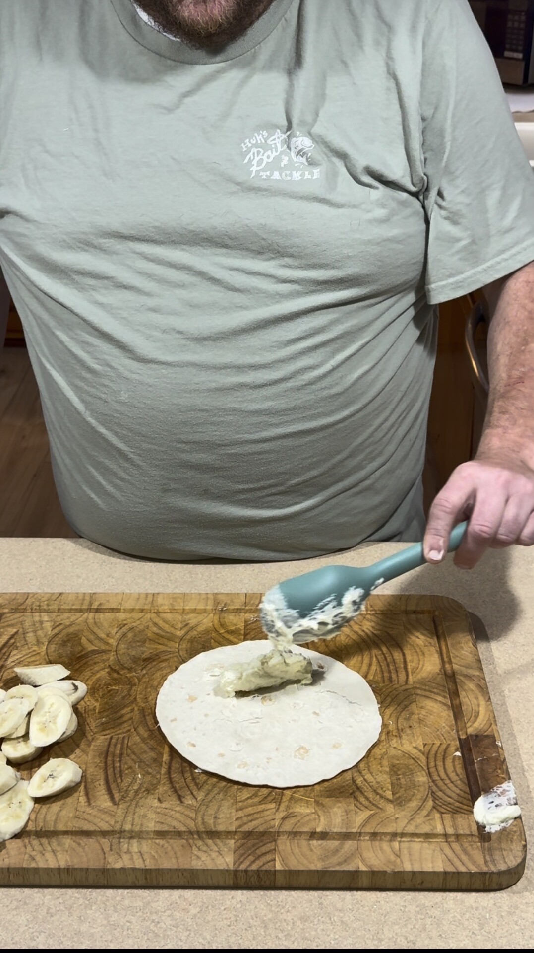 A person spreads a creamy mixture onto a tortilla with a spatula. Sliced bananas are on a wooden cutting board beside the tortilla.
