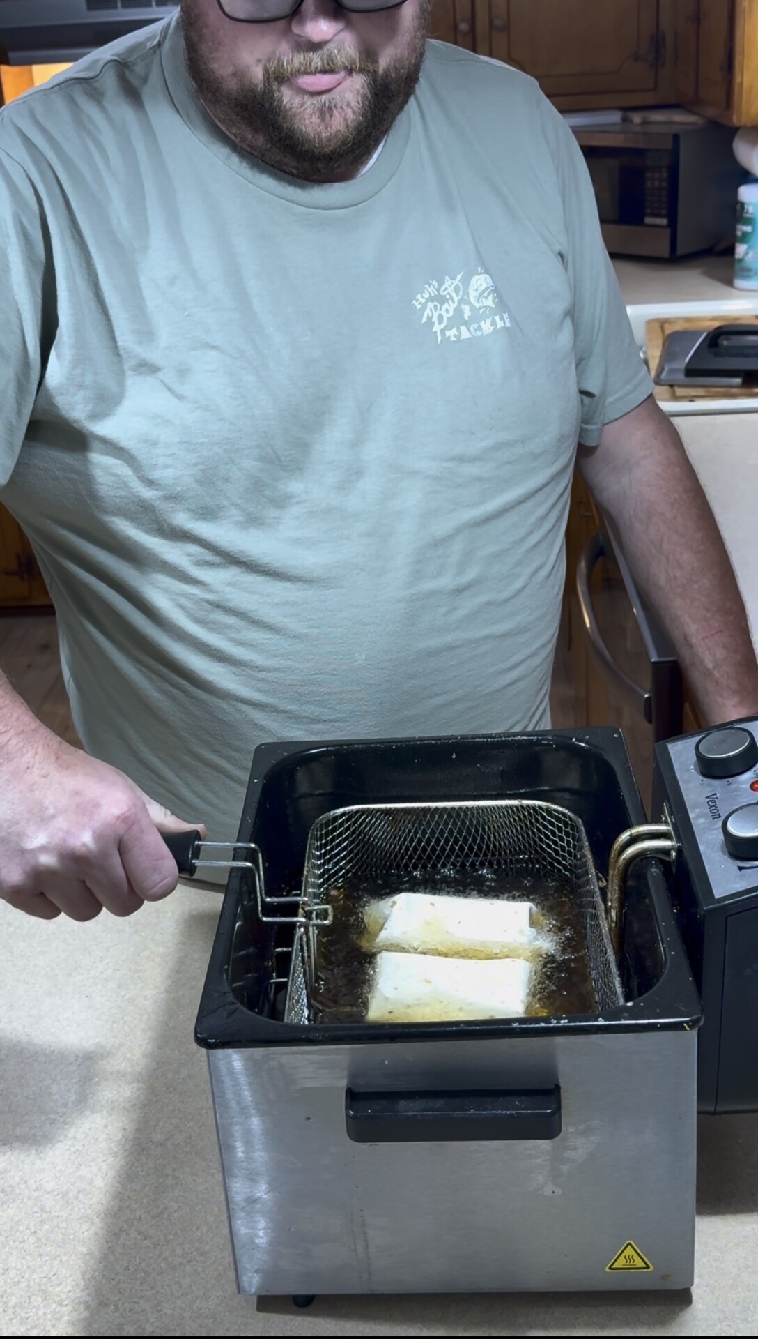 A person in a green shirt holds a basket with food frying in oil inside a deep fryer on a kitchen counter.
