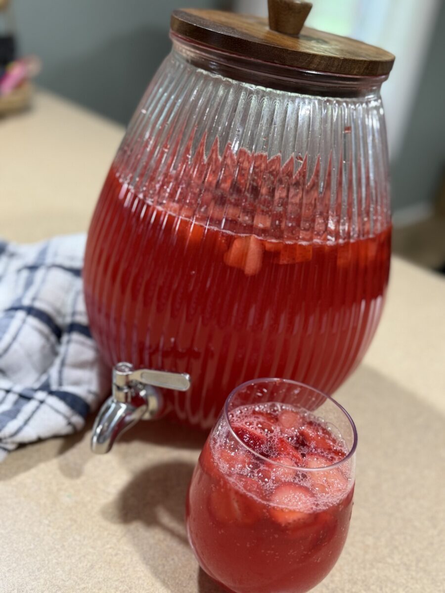 A glass dispenser filled with red fruit punch, with strawberries and ice floating inside. A smaller glass of the punch sits in front, next to a blue and white checkered cloth on a countertop.