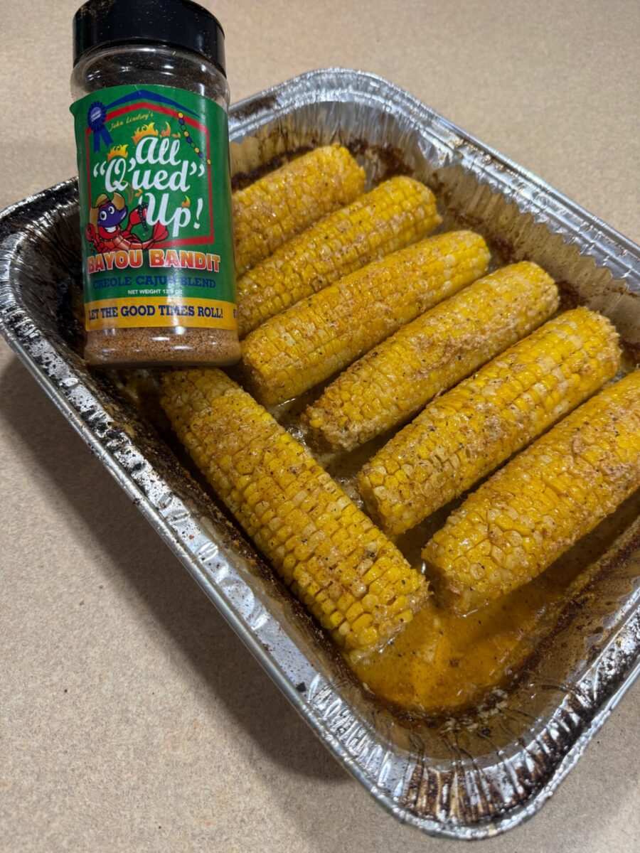 Seasoned corn on the cob in a foil tray with a bottle of All Qued Up Bayou Bandit Cajun blend seasoning resting on the edge.