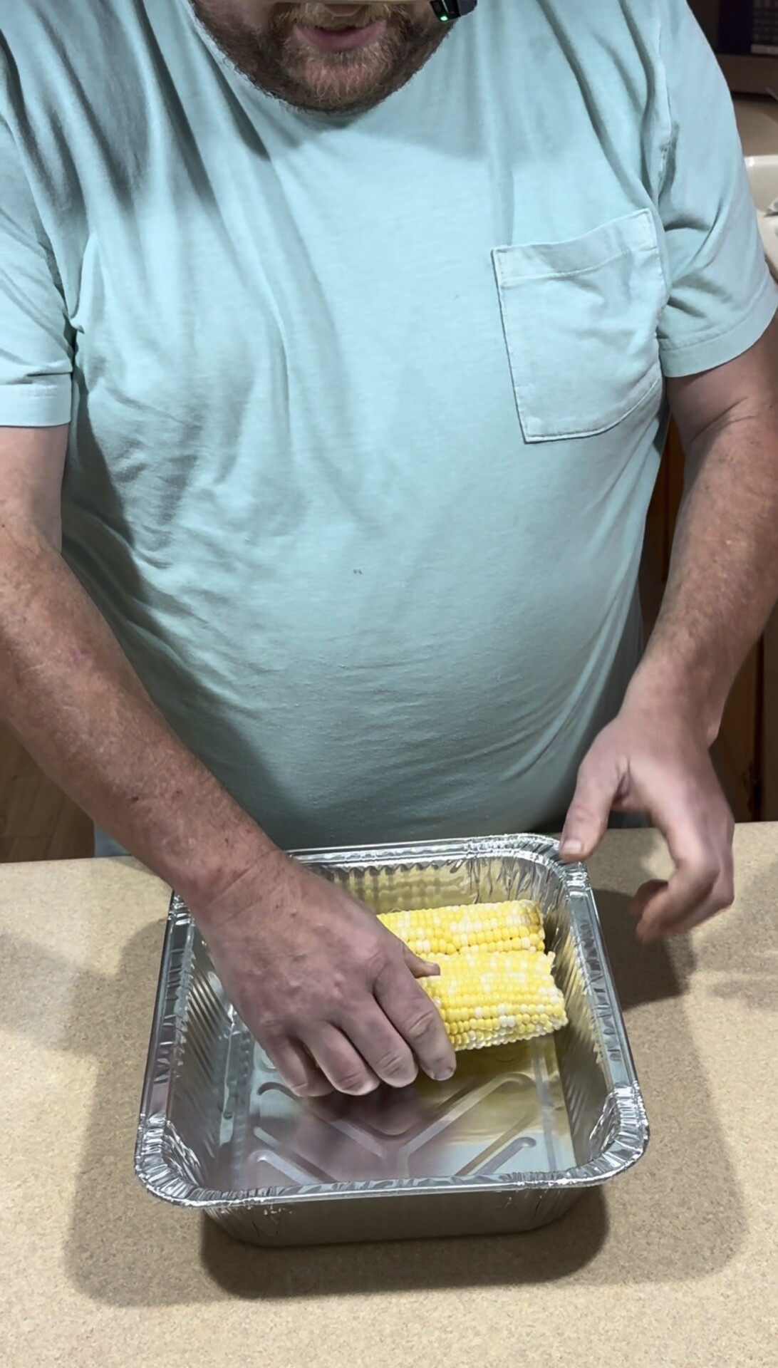 A person in a light blue shirt places several ears of corn into a rectangular aluminum pan on a kitchen counter.