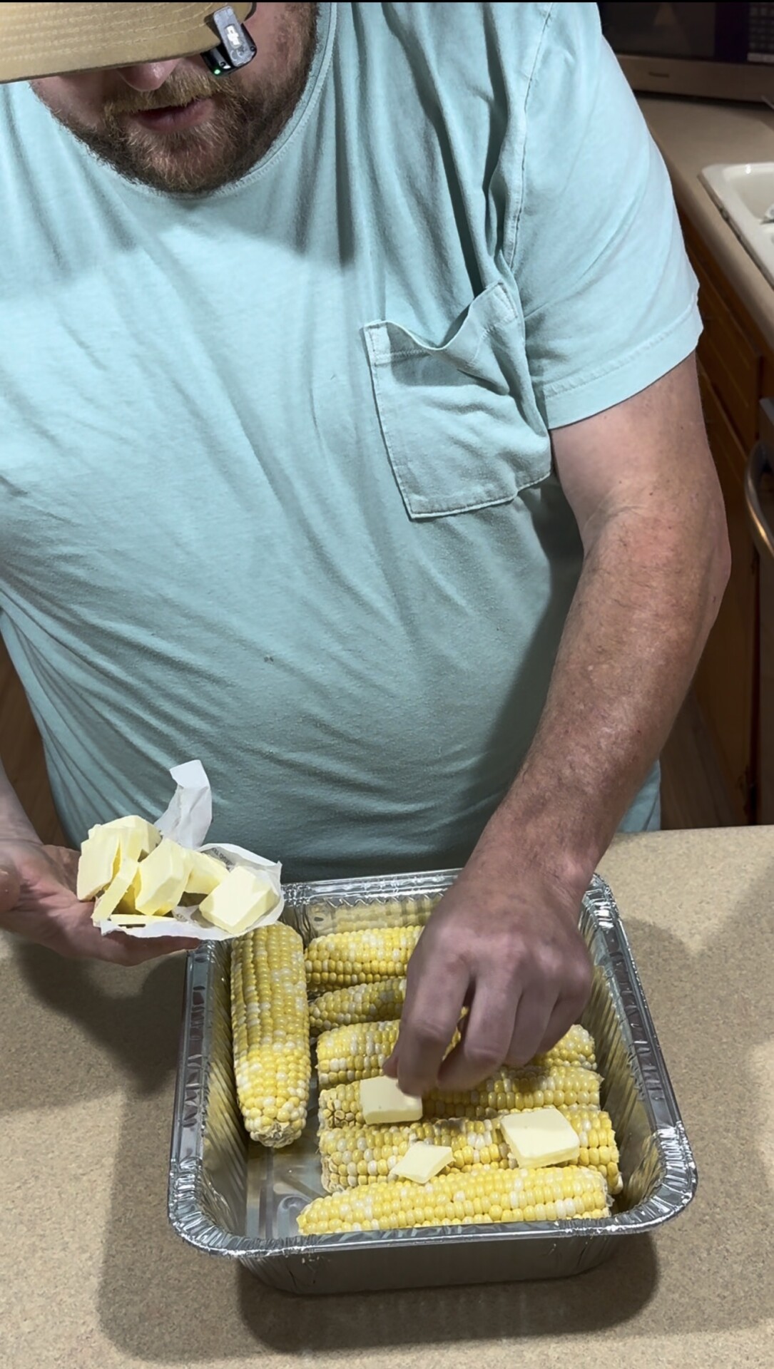 A person in a light blue shirt places slices of butter onto several ears of corn arranged in a foil tray on a kitchen counter.