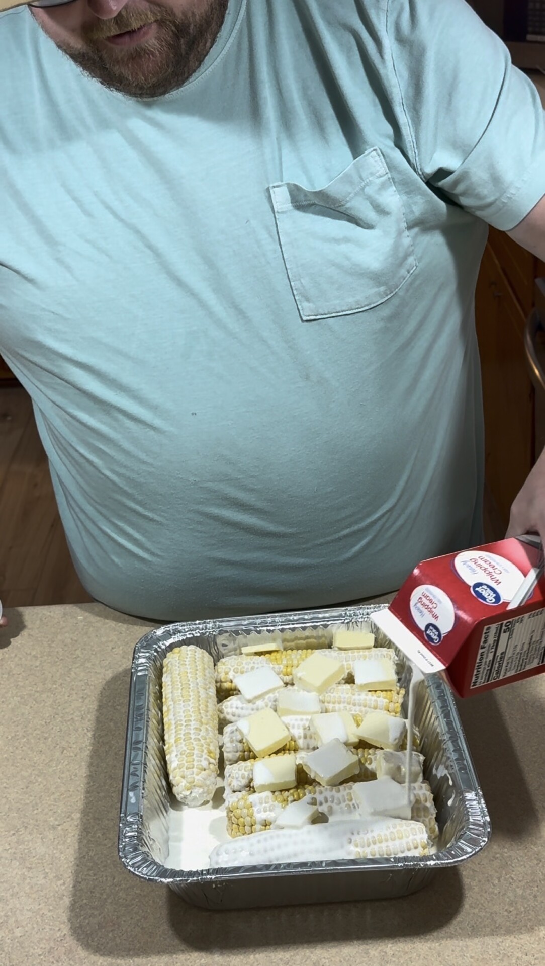 A person in a light blue shirt pours cream from a carton over corn on the cob and butter in a foil pan on a countertop.