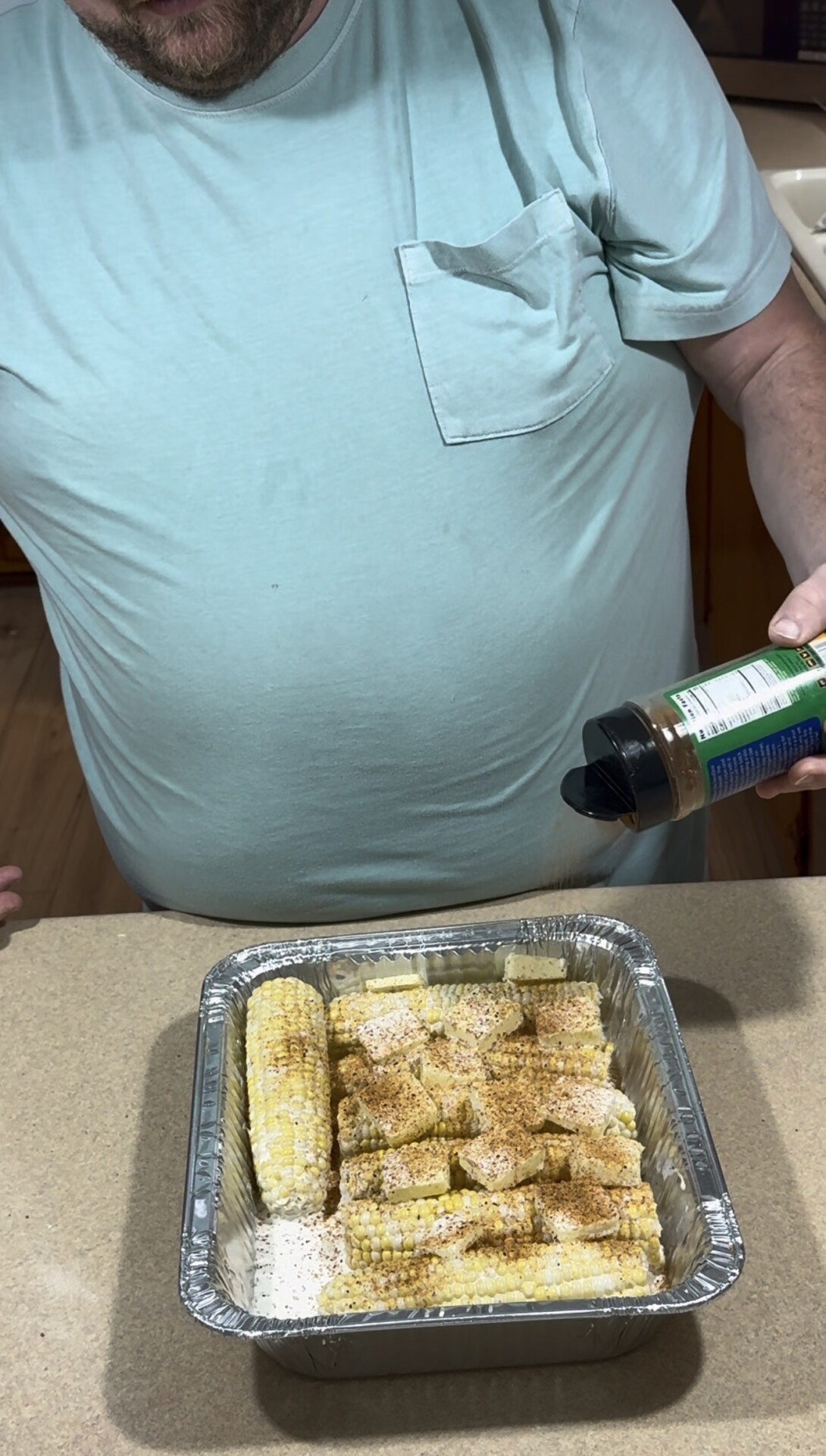 A person in a light blue shirt sprinkles seasoning onto pieces of corn in a foil tray on a countertop.