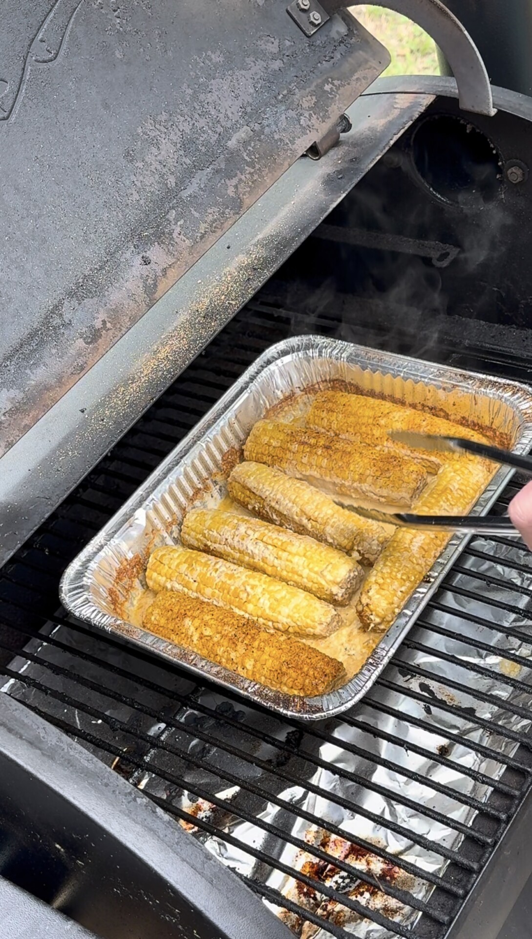 A hand uses tongs to turn corn on the cob in a foil tray on a grill. The corn is coated with seasoning and butter, and steam rises as the grill lid is open.