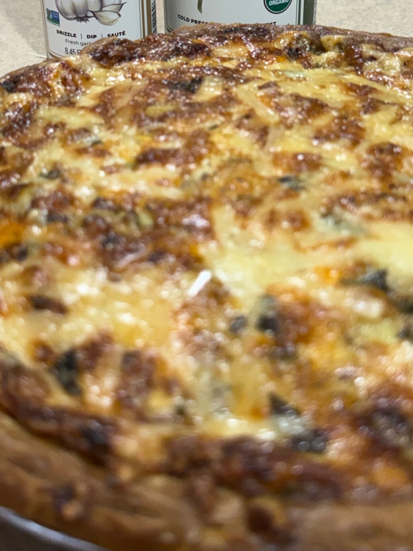 Close-up of a baked quiche with a golden, slightly crispy cheese topping. In the background, there are blurred cans or containers with labels, partially visible.