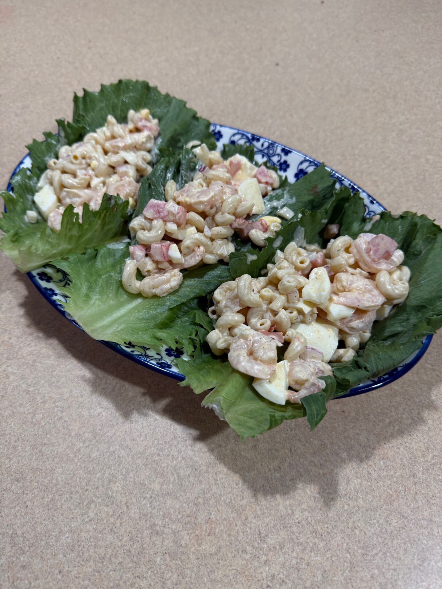 Three lettuce leaves on a blue and white plate, each topped with macaroni salad containing diced ham, egg, and mayonnaise, on a beige counter.