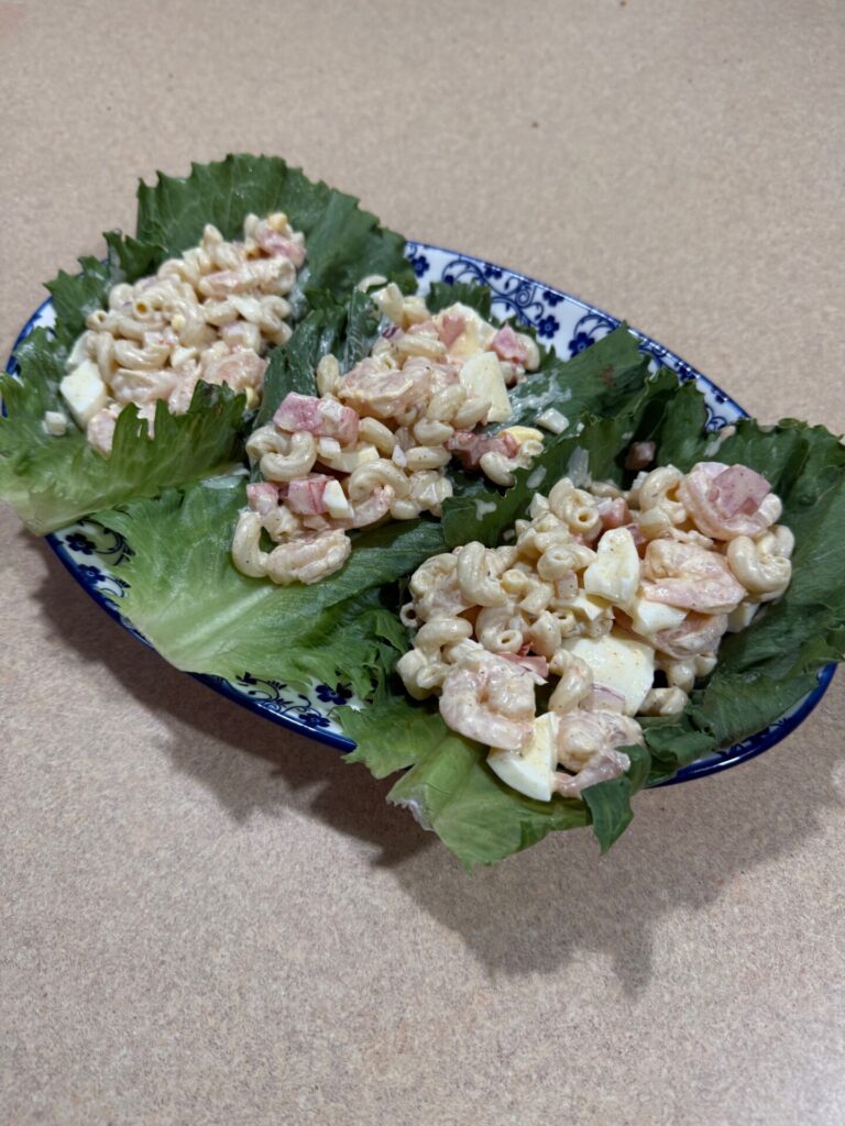 Three lettuce leaves on a blue and white plate, each holding a scoop of macaroni salad with pieces of egg and ham, set on a beige countertop.