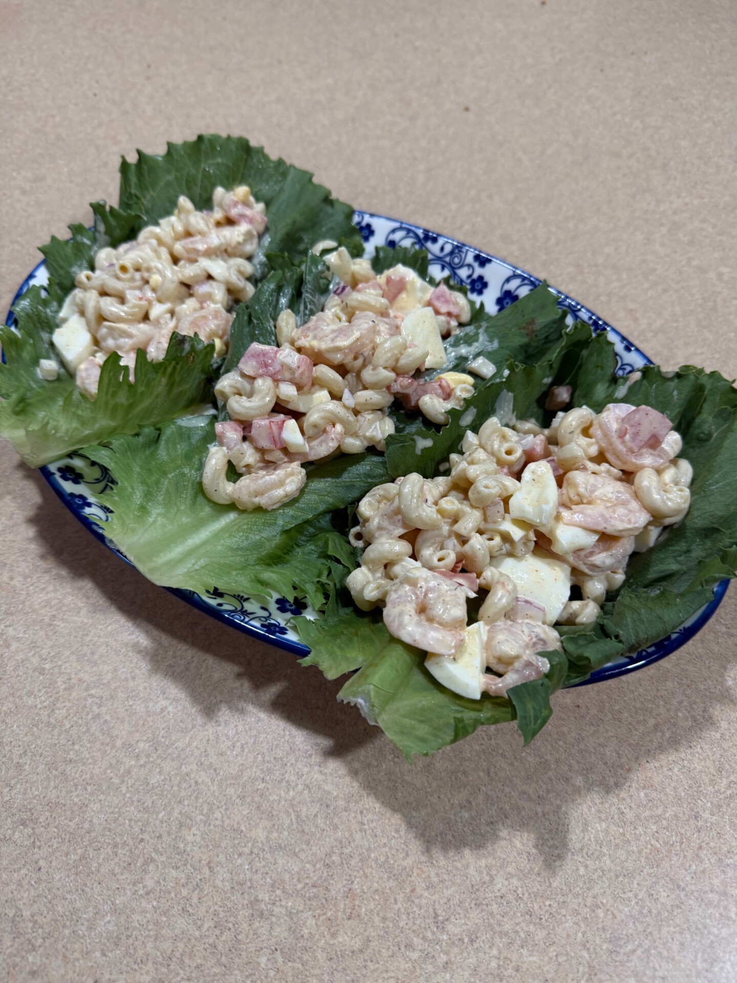 Three lettuce leaves on a blue and white plate, each holding a scoop of macaroni salad with pieces of egg and ham, set on a beige countertop.