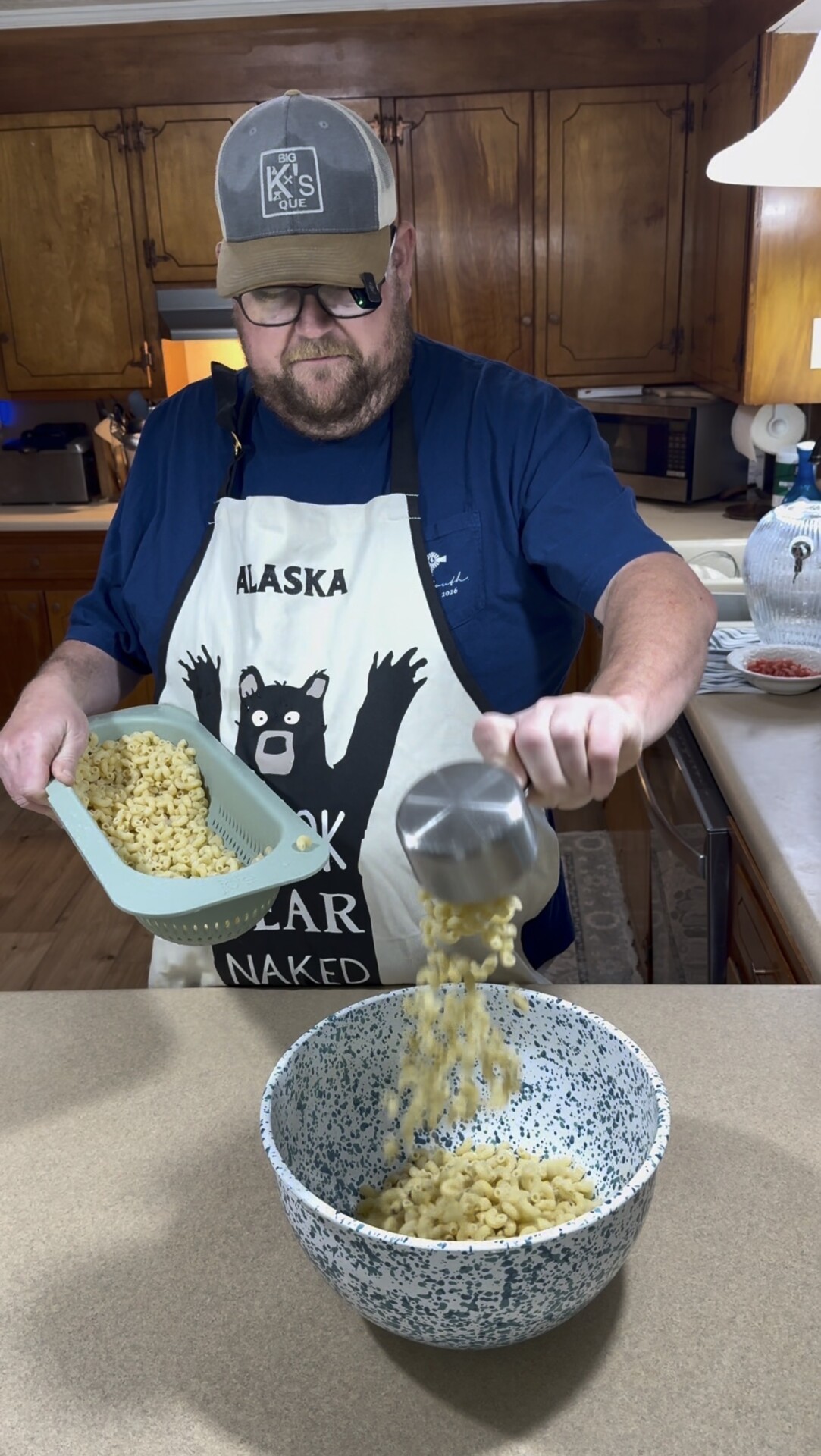 A man wearing an apron with a bear on it pours cooked macaroni from a cup into a speckled mixing bowl in a kitchen.