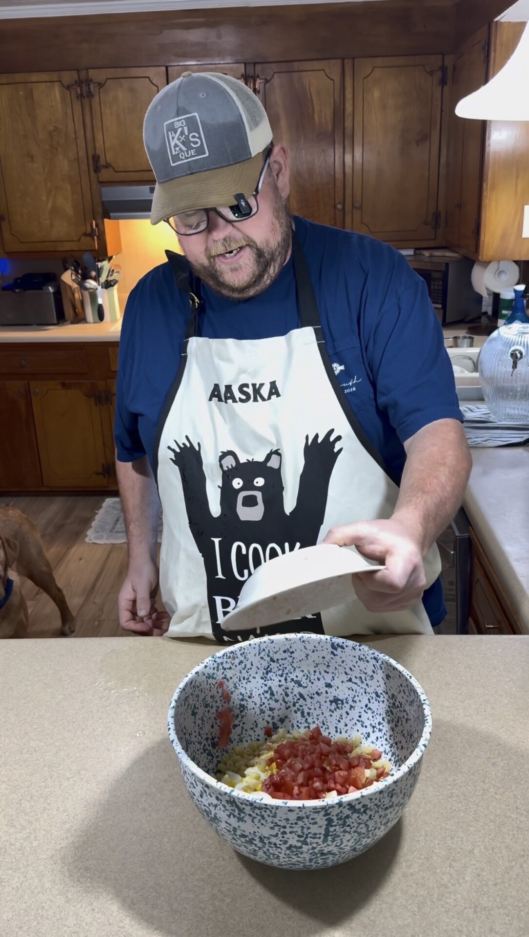 A man wearing an Alaska I Cook bear apron and cap pours chopped vegetables into a speckled bowl in a kitchen. A brown dog stands in the background, partially visible.