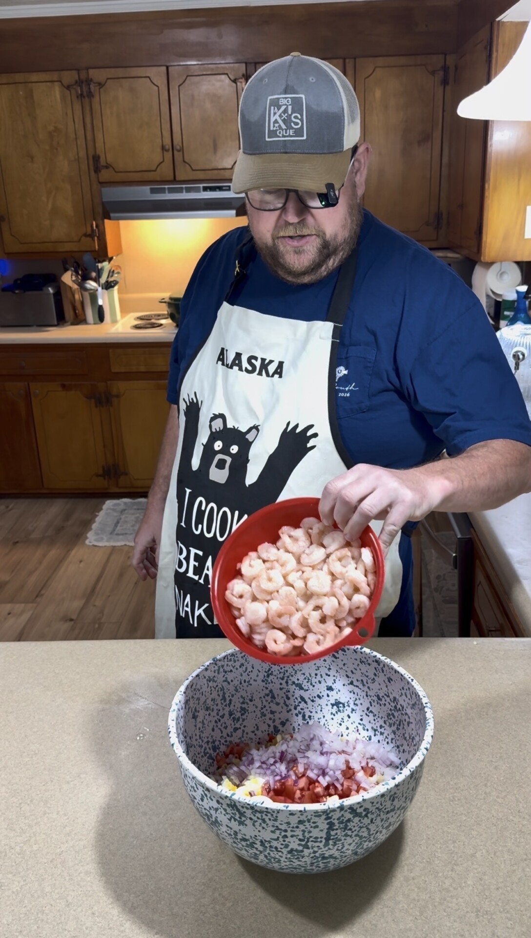 A person in an “Alaska I cook bear naked” apron pours shrimp from a red bowl into a large speckled mixing bowl filled with chopped vegetables in a kitchen.