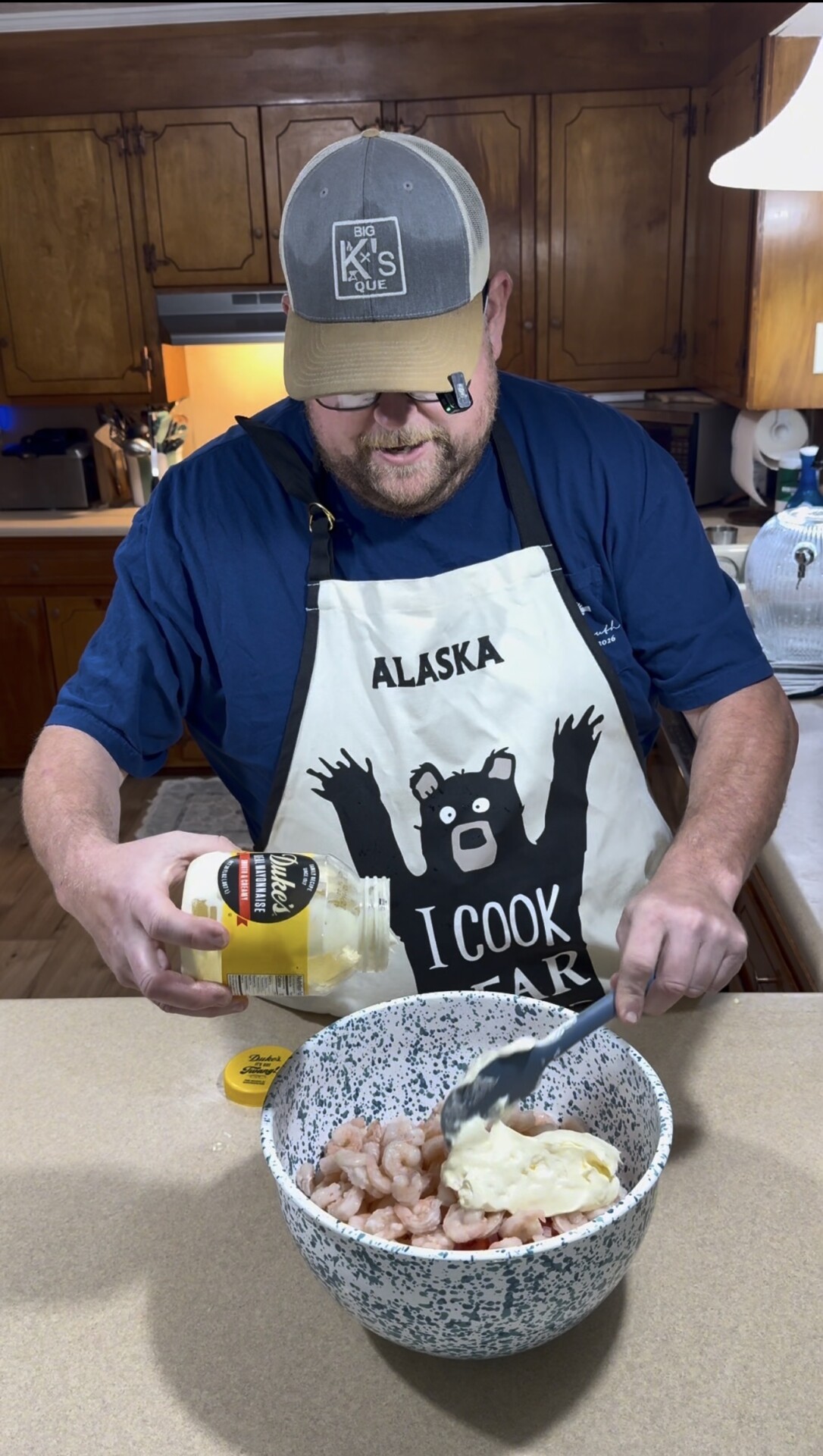 A man wearing an “ALASKA I COOK BEAR” apron pours mayonnaise into a large bowl of shrimp and vegetables in a kitchen. He uses a spoon to mix the ingredients while smiling.