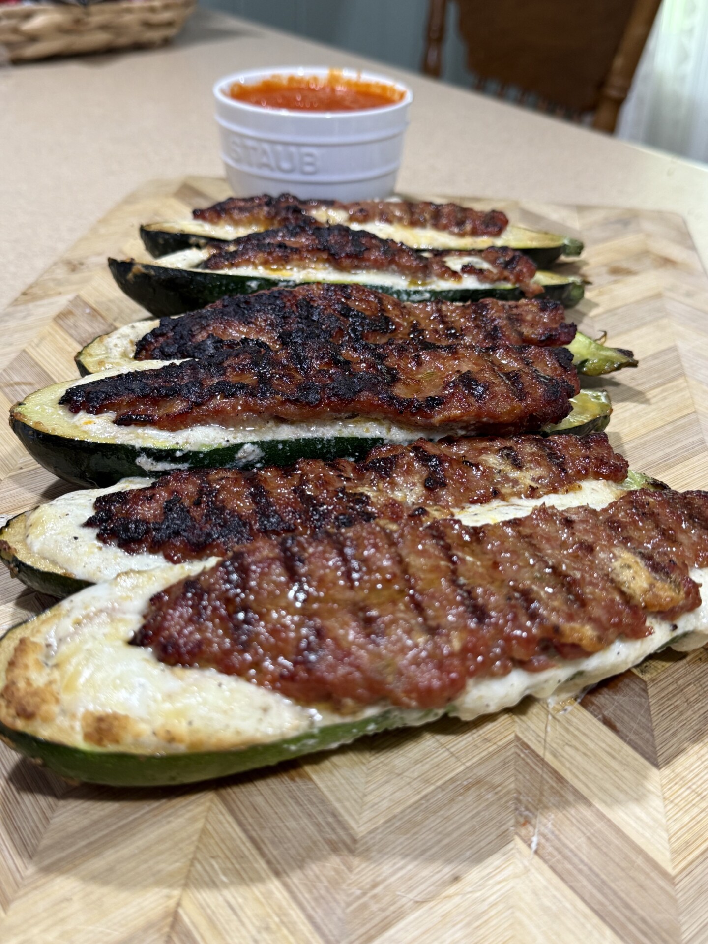 Four grilled, stuffed zucchini halves with browned tops are arranged on a wooden cutting board. A small white ramekin filled with red sauce is in the background.