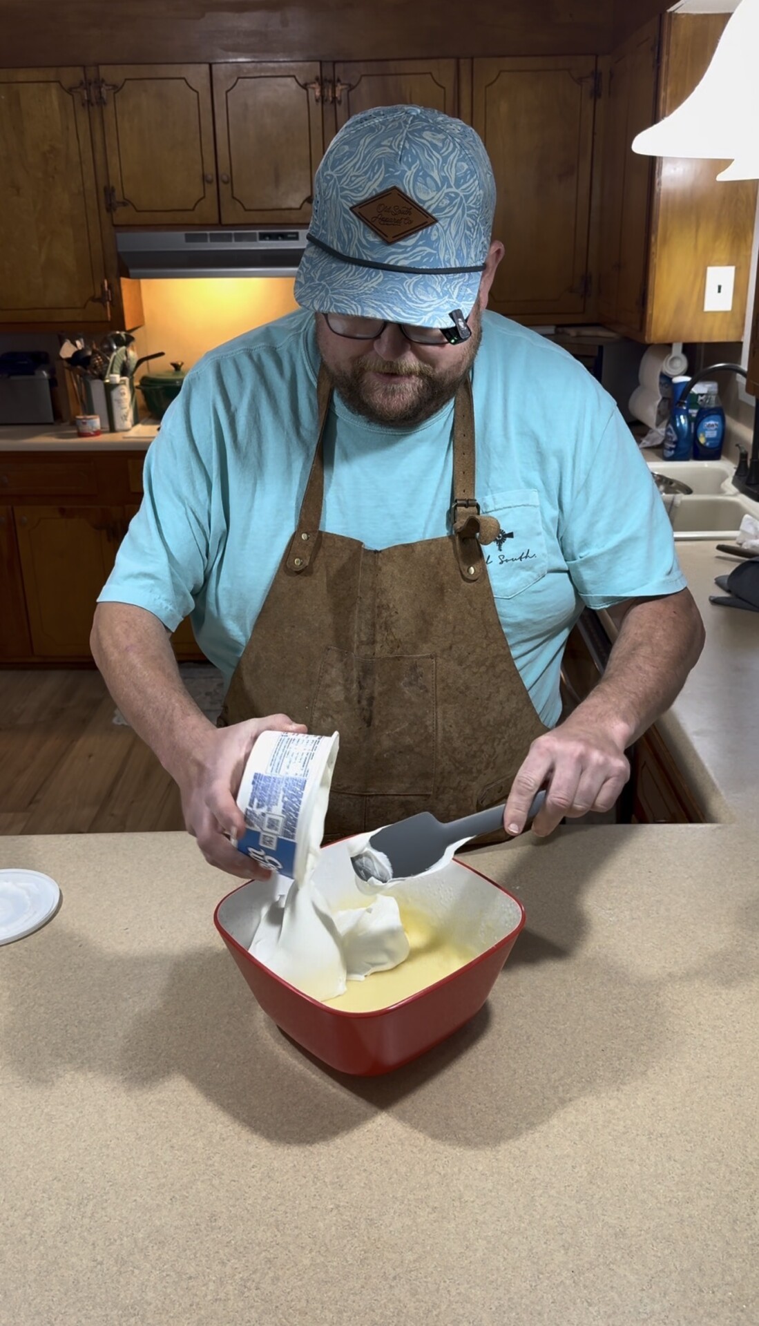 A man wearing a blue hat and apron stands in a kitchen, scooping whipped topping from a container into a red bowl on the counter.