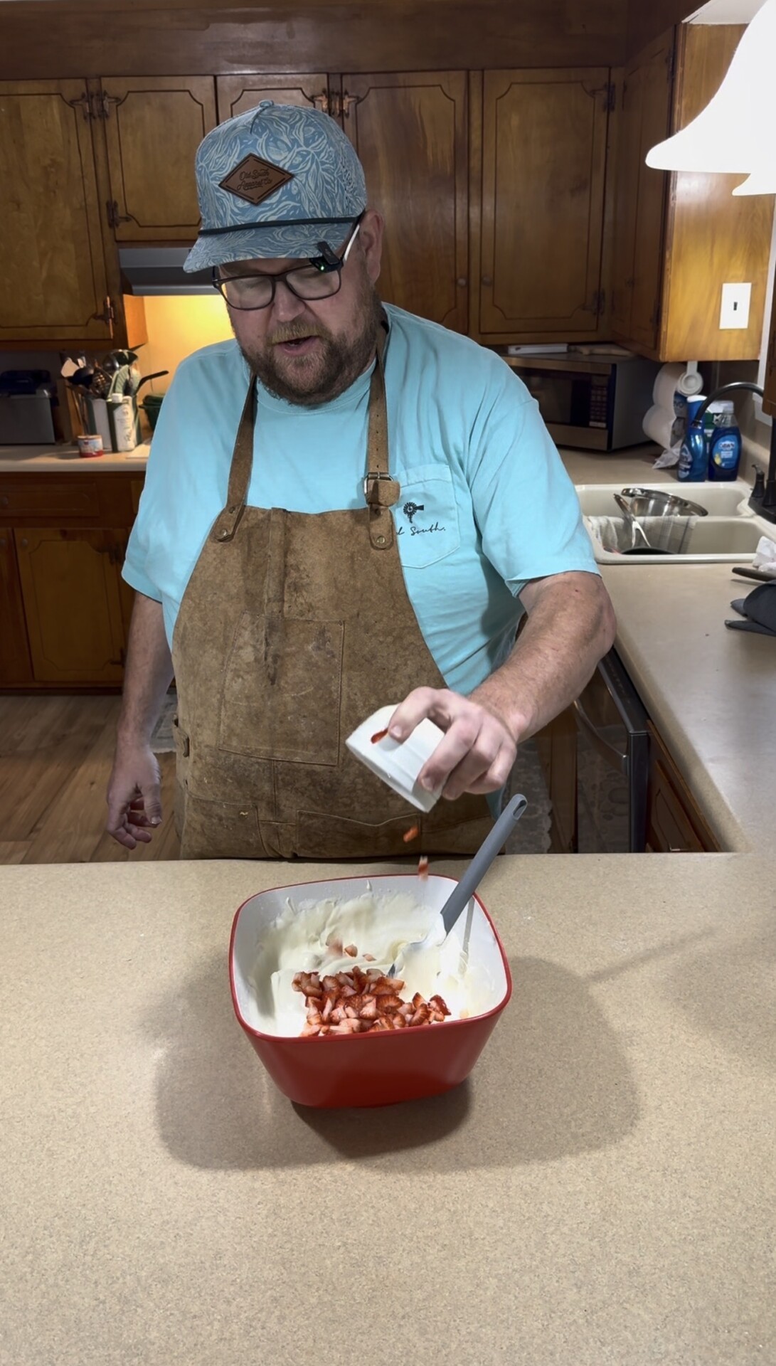 A man in a blue shirt and brown apron sprinkles chopped strawberries from a cup into a mixing bowl of cream in a kitchen.