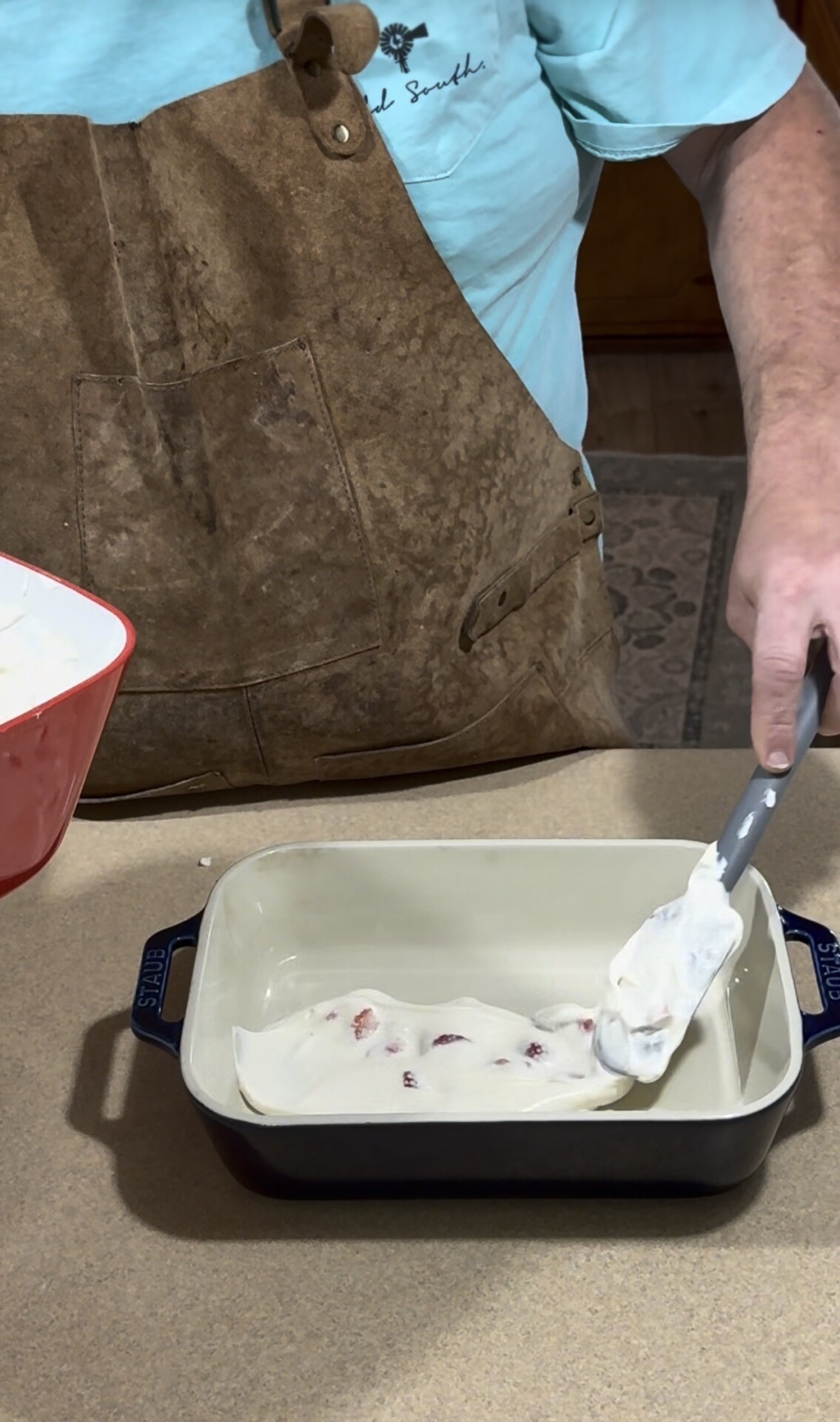 A person in a brown apron spreads a creamy white mixture with a spatula into a rectangular baking dish on a kitchen counter.